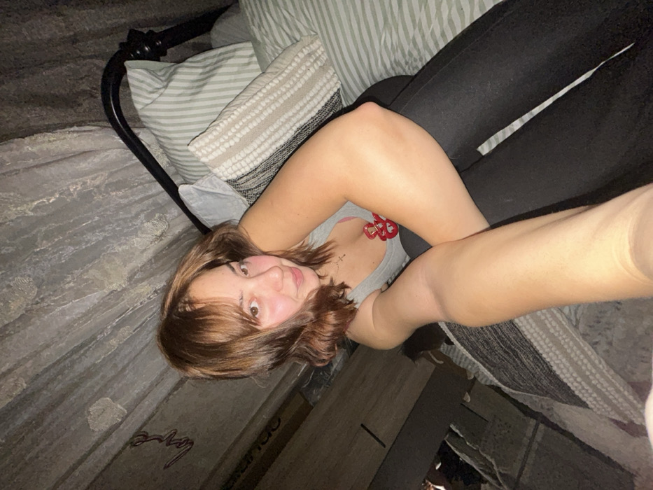 A young woman with short brown hair lies on a bed. She wears a gray tank top and black pants. Her arm is outstretched, suggesting she is taking a selfie. Pillows with stripes and texture are behind her head. The bed has a black metal frame. The wall near the bed is light gray with a faded word. The lighting is dim, casting shadows in the scene.
