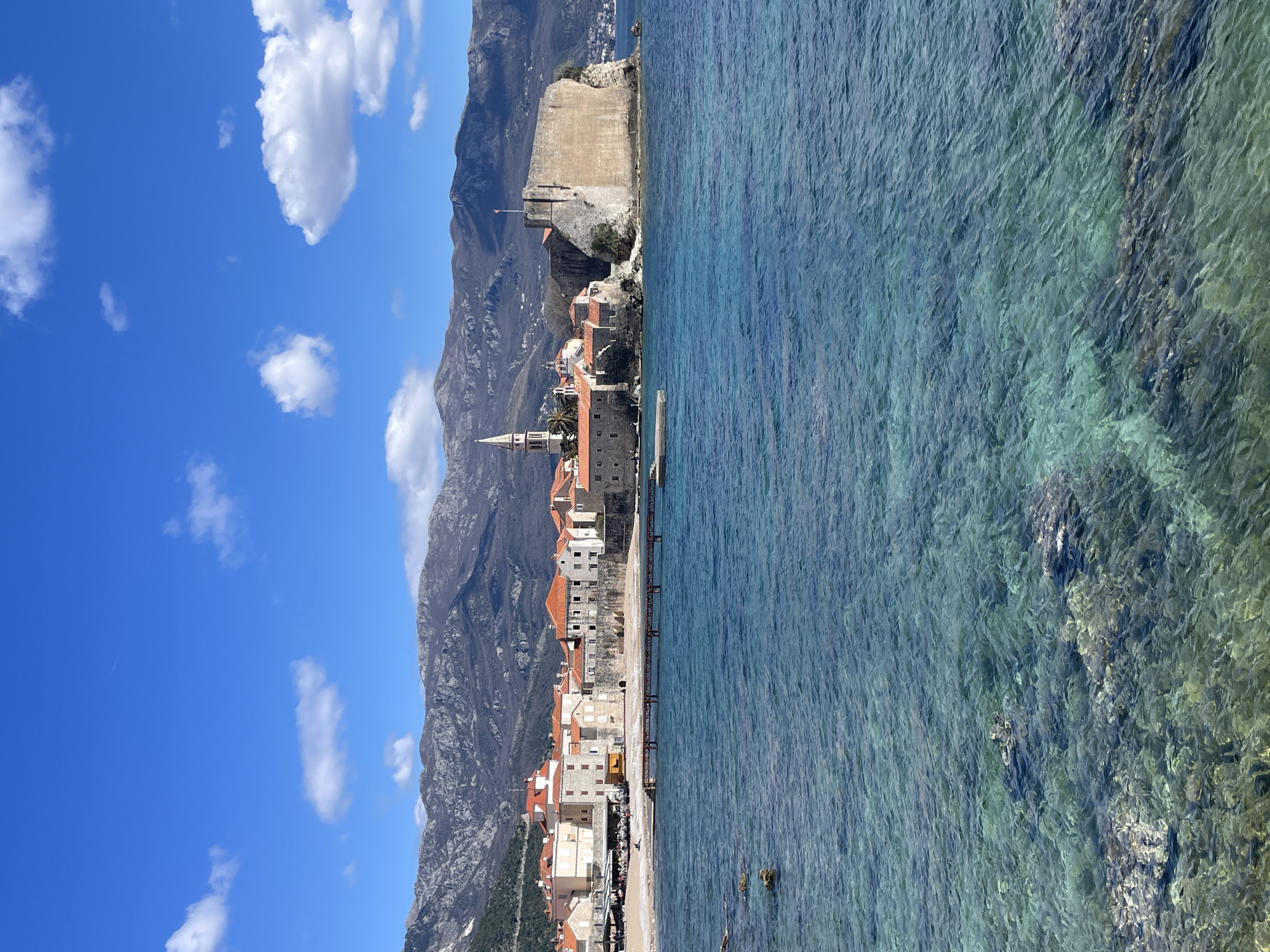 The image displays a coastal scene of Budva Old Town (Stari Grad) in Montenegro. The foreground features clear, light blue-green seawater with visible underwater rock formations and a rippled surface. In the midground, the fortified peninsula of Budva Old Town extends into the water from left to right. It comprises numerous multi-story stone buildings with off-white facades and terracotta-tiled roofs. A tall, light-colored church steeple with a pointed cap is prominent on the left side of the old town structure. To the right, a large, robust stone fortification with high walls and battlements forms the seaward edge of the peninsula, topped by a flagpole bearing a small orange flag. Adjacent to the left side of the old town, a narrow strip of sandy or pebbly beach is visible, where several people are seated at outdoor tables under umbrellas, indicating a commercial establishment. A brown or reddish pier extends from this beach area into the water. A single small, white boat is moored near the pier. The background consists of a range of rugged, grey-brown mountains with visible rocky outcrops and some sparse vegetation, stretching across the horizon under a bright, clear blue sky containing scattered white cumulus clouds and a faint airplane contrail.