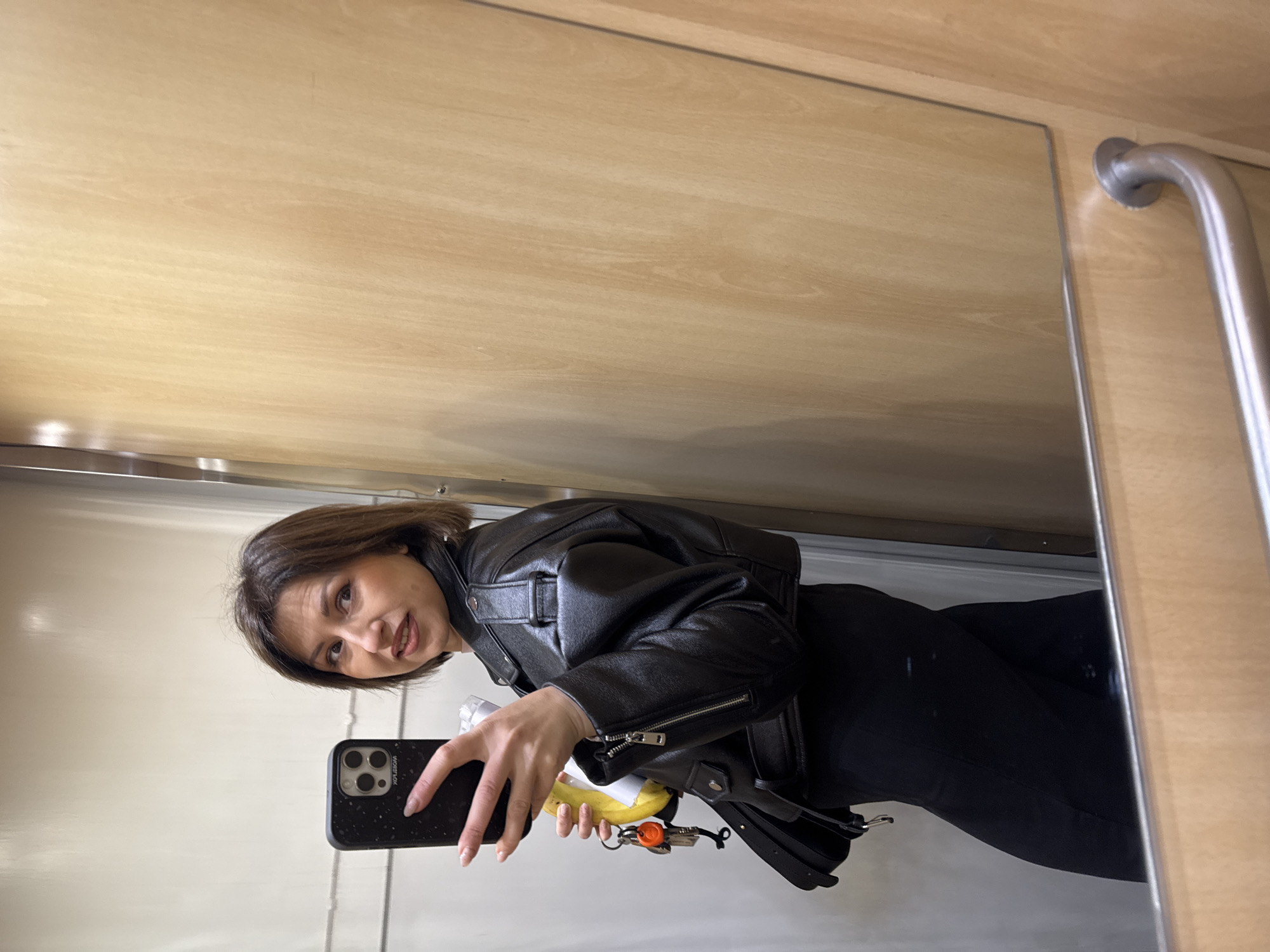 The image shows a woman taking a selfie in a mirrored elevator. The woman has short brown hair and is wearing a black leather jacket and black pants. She is holding a black smartphone in her right hand. In her left hand, she's holding a banana and a set of keys. The elevator has light brown wood paneling and a chrome handrail. The location is not identifiable as Sofia, Bulgaria.
