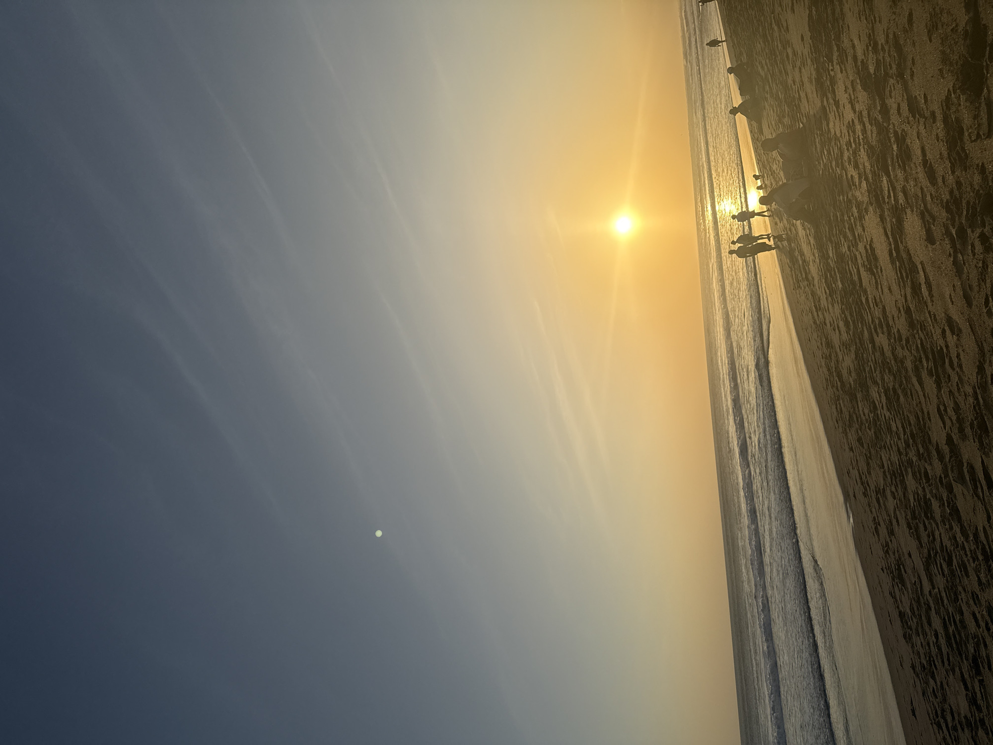 The image depicts a sunset on a sandy beach in North Kuta, Indonesia. The sky transitions from a deep blue at the top to a soft yellow and orange near the horizon where the sun is setting. Wispy clouds streak across the sky, catching the golden light. The sun, a bright, radiant orb, casts a shimmering reflection on the ocean. Gentle waves roll onto the shore, creating a foamy white line. Silhouettes of people are scattered across the beach and near the water's edge. Some are standing and walking, while others are sitting on the sand, seemingly enjoying the sunset. The sand itself is dark and textured with footprints and natural patterns. The overall scene conveys a sense of tranquility and the end of a day at the beach.
