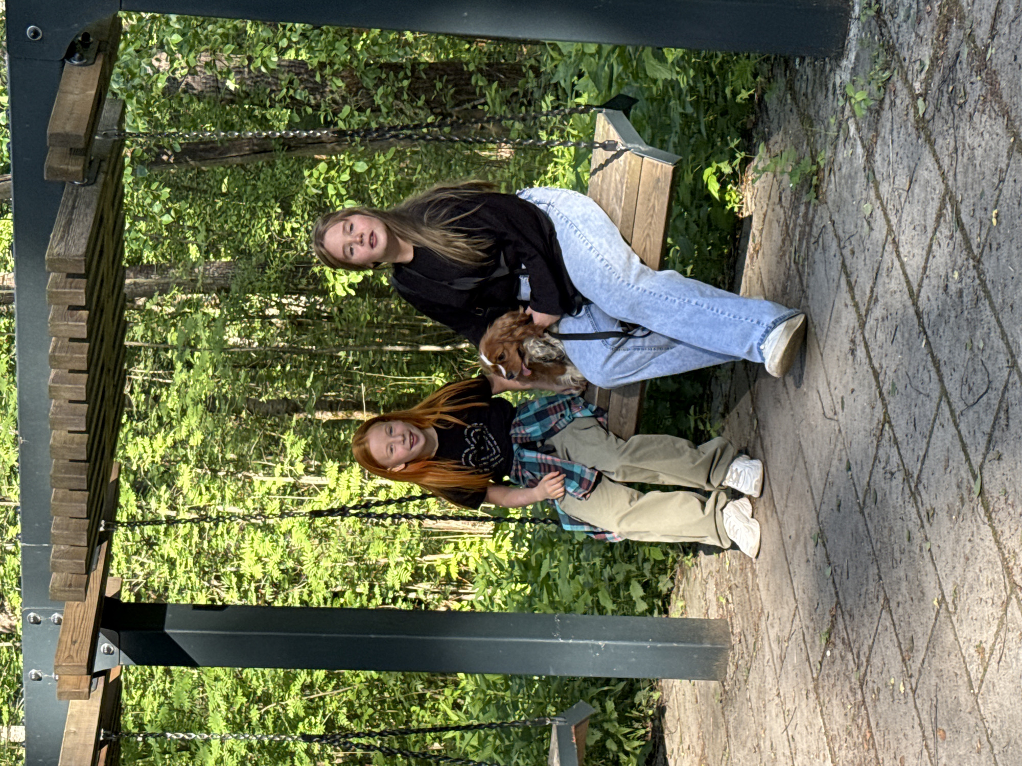 Two young women are sitting on a wooden swing under a dark metal frame structure. One woman, with long brown hair, is wearing a black top and light blue jeans, and she is holding a brown and white dog. The other woman, with long red hair, is wearing a black top with a heart design, a blue and green flannel tied around her waist, and khaki pants. The swing is located in a park-like setting with lush green trees in the background, and the ground is paved with gray stone tiles. The image is rotated counter-clockwise. There is no indication of location in Veliky Novgorod, Russia.