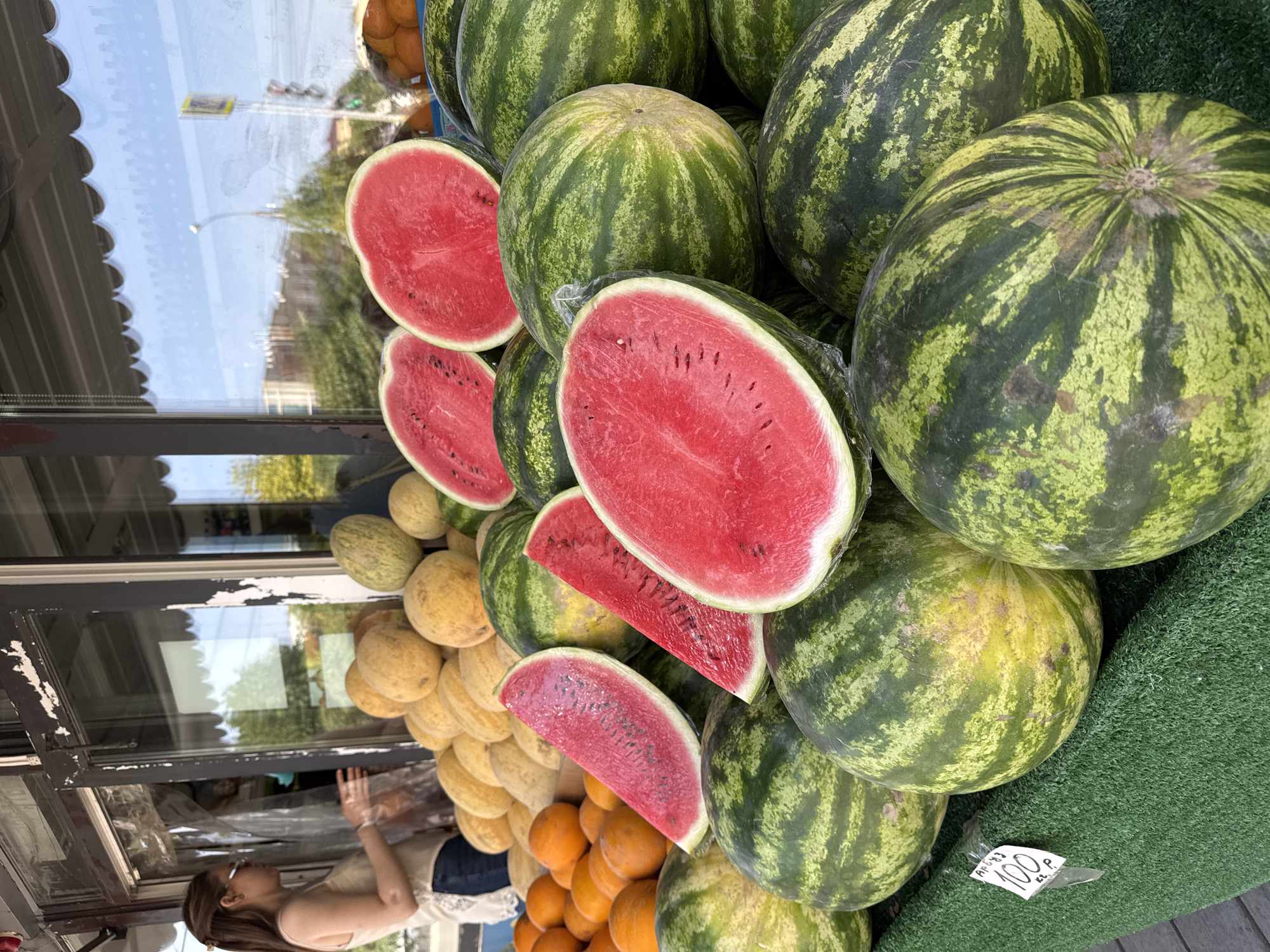 A medium shot captures a street-side fruit stand in Novosibirsk, Russia, on a bright, sunny day. In the foreground, a large display of watermelons is arranged, with several cut in half to reveal their vibrant red flesh. Behind the watermelons, there are stacks of yellow melons and oranges. To the left of the frame, a young woman wearing sunglasses, a light-colored tank top, and denim jeans is interacting with a vendor behind a plastic curtain. The background shows a building facade with glass windows reflecting the sky and city. The overall scene depicts a typical urban market environment, showcasing a variety of fresh produce.