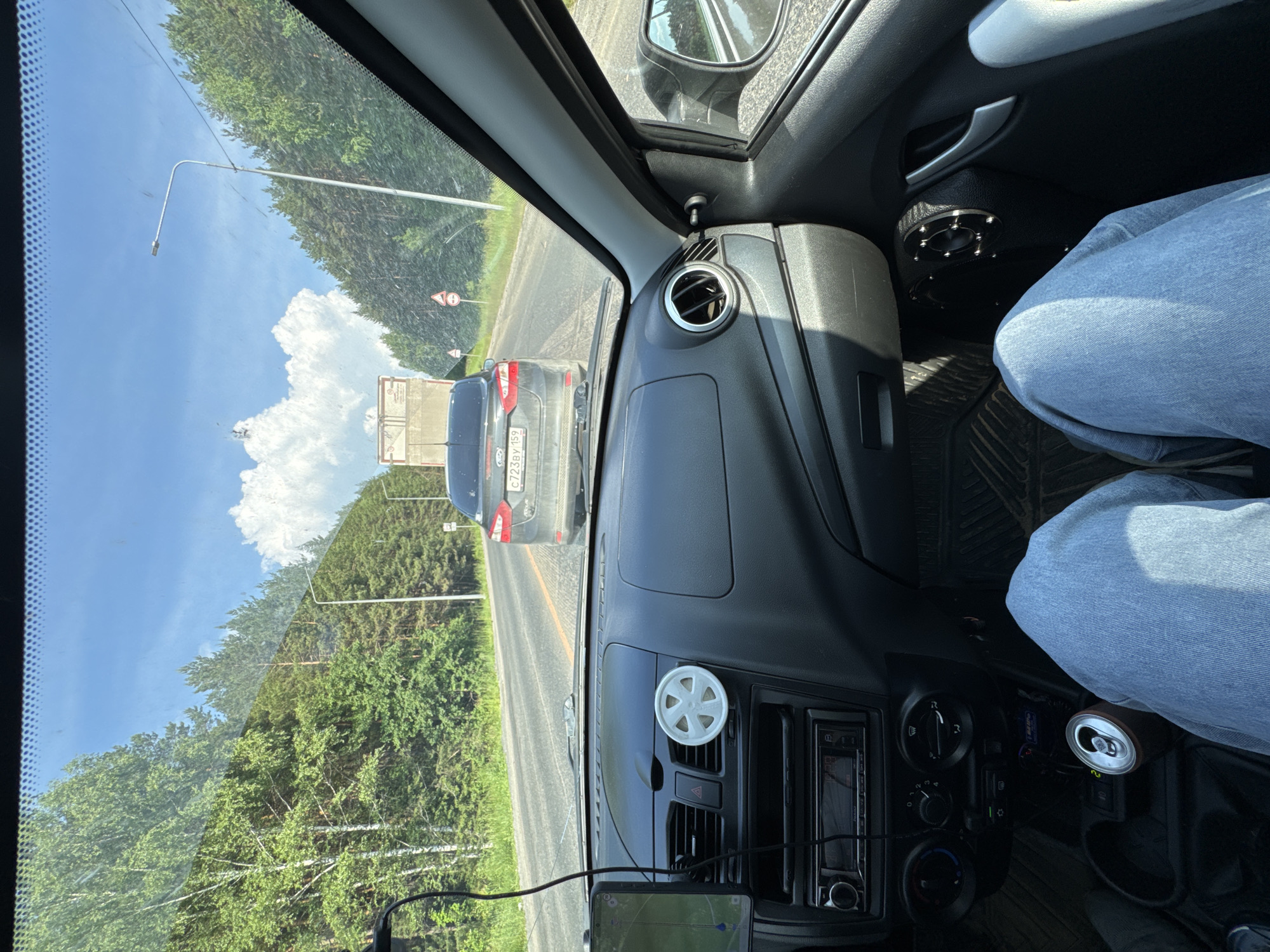 The image depicts a view from the driver's seat of a car traveling on a road lined with a dense forest of birch and pine trees under a bright, cloudy sky. The road appears to be a two-lane highway. Ahead, a dark gray Ford sedan is visible, its license plate reading "C723BY 159". Further ahead, a white truck with a beige cargo area is moving in the same direction. Several road signs are present along the roadside, including what appear to be speed limit or cautionary signs.
Inside the car, the dashboard is visible, featuring an air conditioning vent with a white decorative element, a car stereo, and a navigation device mounted on the left. The driver's legs, clad in blue jeans, are in the foreground. A can of soda is placed in a cup holder. The overall environment suggests a daytime journey through a rural or semi-rural area, possibly on a highway leading to or from Pesterevo, Russia, given the context.