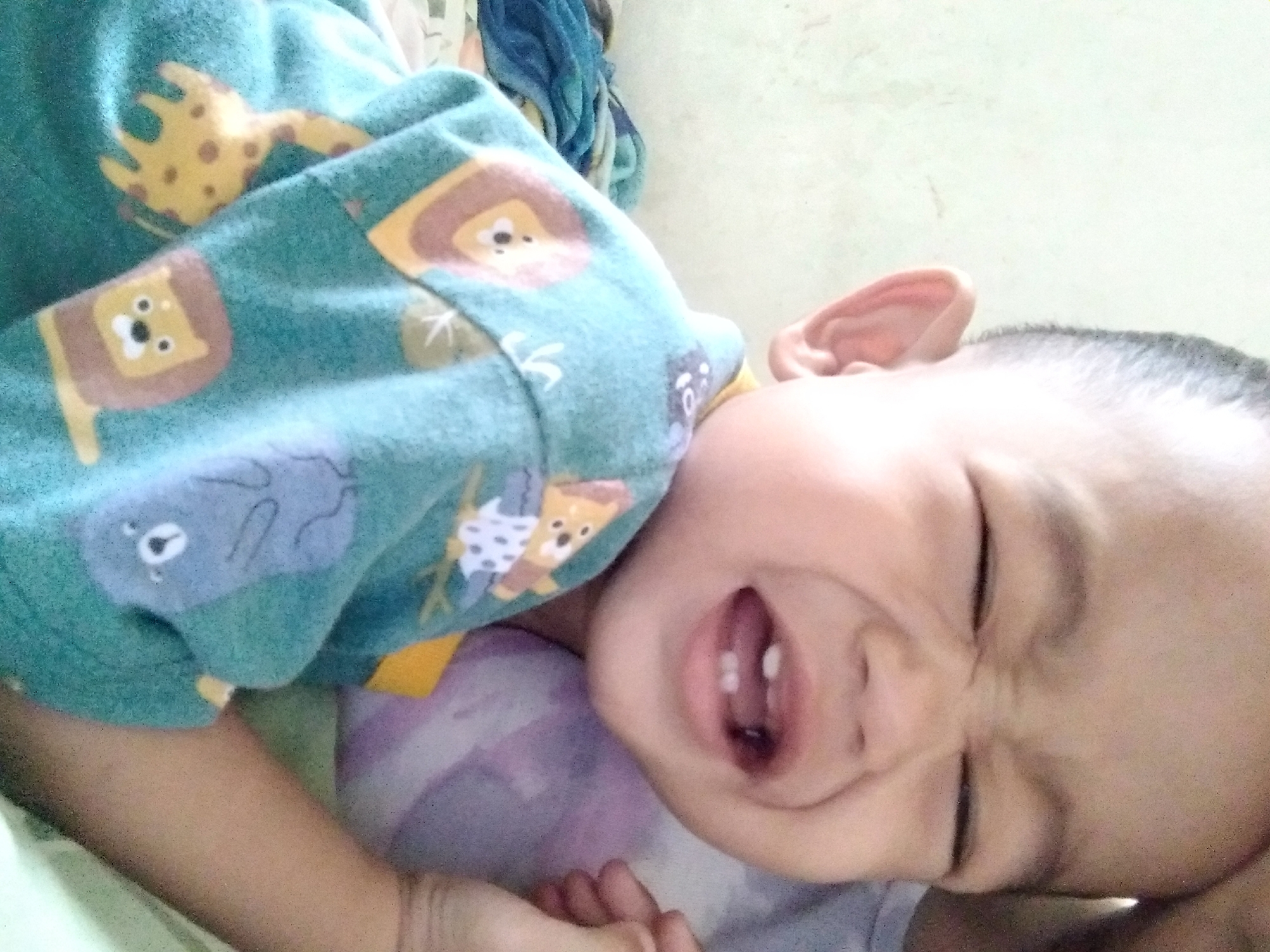 A close-up, over-the-shoulder shot captures a young child, likely a toddler, with eyes squeezed shut and mouth open in an expression of apparent distress or laughter. The child is wearing a teal-colored shirt adorned with cartoon jungle animals, including lions, giraffes, and what appear to be elephants or koalas. The background is softly lit and out of focus, suggesting an indoor setting. Part of a purple object, possibly a pillow or toy, is visible behind the child's shoulder. The scene appears to be a personal photograph, potentially taken in a home environment in Pekanbaru, Indonesia, given no other context.