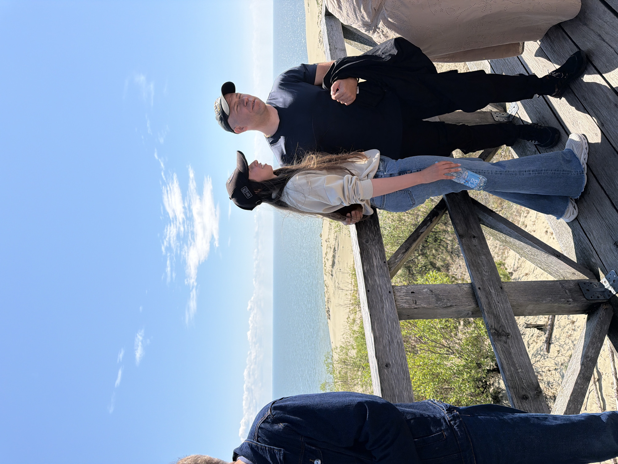 In Morskoye, Russia, on a wooden boardwalk overlooking a sandy dune landscape and the sea, a man and a young woman are standing. The man, wearing a dark t-shirt and a baseball cap, holds a black jacket in his right hand. The young woman, wearing a light-colored sweatshirt, jeans, and a baseball cap, is holding a bottle of water. To the left, the back of another person wearing a denim jacket is visible. Another person, mostly obscured, is visible on the far right. The sky is blue with scattered clouds, and the sea is calm.