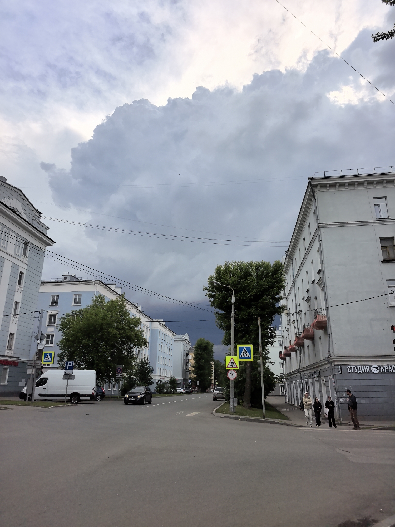 An urban street scene unfolds under a dramatic, heavily clouded sky, suggesting impending rain or a recent storm. The street, possibly an intersection or a bend, is lined with multi-story buildings and trees.
On the left, a light blue building with white trim stands prominently, and a white cargo van is parked nearby. Further down the street, a dark sedan is driving. Overhead power lines crisscross the upper part of the frame.
On the right side of the street, a light grey or white building features several small, decorative balconies. The ground floor of this building hosts a business with a sign reading "СТУДИЯ КРАСЫ," which translates to "BEAUTY STUDIO." In front of this building, four individuals are visible on the sidewalk. Three are standing together, seemingly conversing or waiting, while a fourth person on the far right appears to be looking down or adjusting something.
Several road signs are present: yellow pedestrian crossing signs, a blue 'P' indicating parking, and a red-bordered circular sign displaying "40" for a speed limit. Streetlights and traffic signals are also visible, with the traffic light on the right showing a red signal. The time of day appears to be daytime, possibly late afternoon, with the diffused light from the dense cloud cover creating a somber, muted atmosphere.