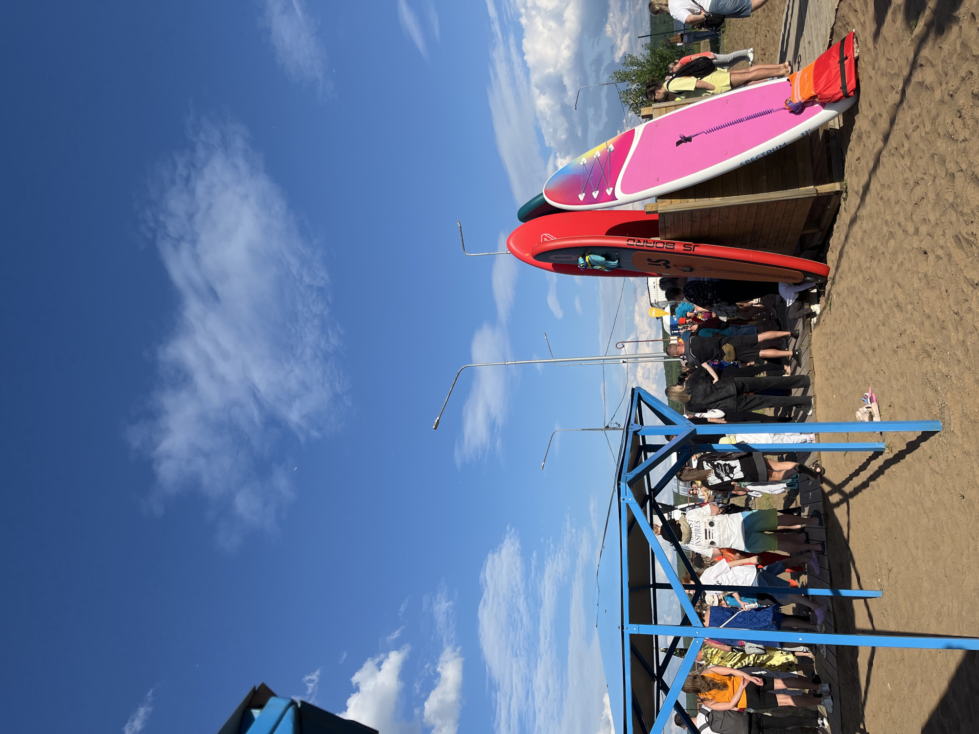 The image depicts a sunny day with a bright blue sky and scattered white clouds. In the foreground, a blue metal structure with a roof covers a sandy area where several people are gathered. To the right, three paddleboards are propped up against a wooden stand. The paddleboards are red, green, and pink with colorful designs. A crowd of people is visible behind the blue structure and near the paddleboards, some appearing to be waiting. In the background, there are streetlights, trees, and a fence. The scene appears to be an outdoor recreational area, likely a beach or a waterfront, possibly in the city of Chaikovsky, Russia, given the context.