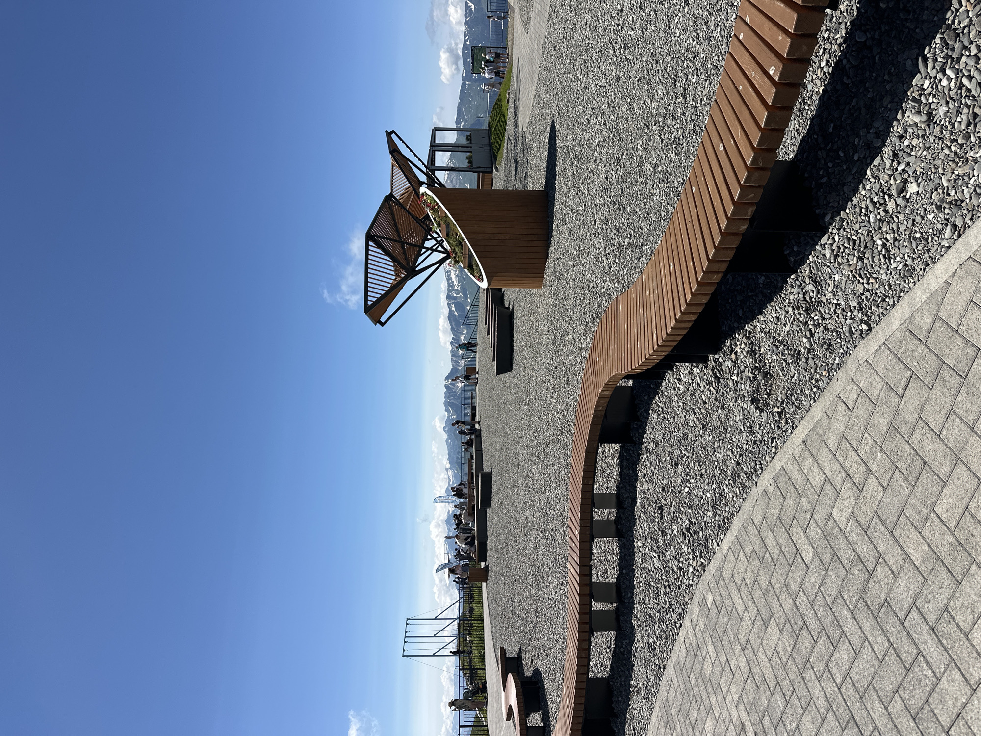 This is an outdoor, high-angle, eye-level shot of a scenic overlook in Estosadok, Russia. The foreground features a winding, wooden bench set on a gravel path. Beyond the bench, more gravel covers the ground, with a central, abstract wooden sculpture housing a planter filled with flowers. Further back, a group of people are scattered around the overlook, some standing, others seated. In the background, a picturesque mountain range with snow-capped peaks stretches across the horizon under a clear blue sky with a few wispy clouds. A suspension bridge-like structure is visible to the left. The overall impression is one of a well-maintained tourist attraction designed to showcase the natural beauty of the area.