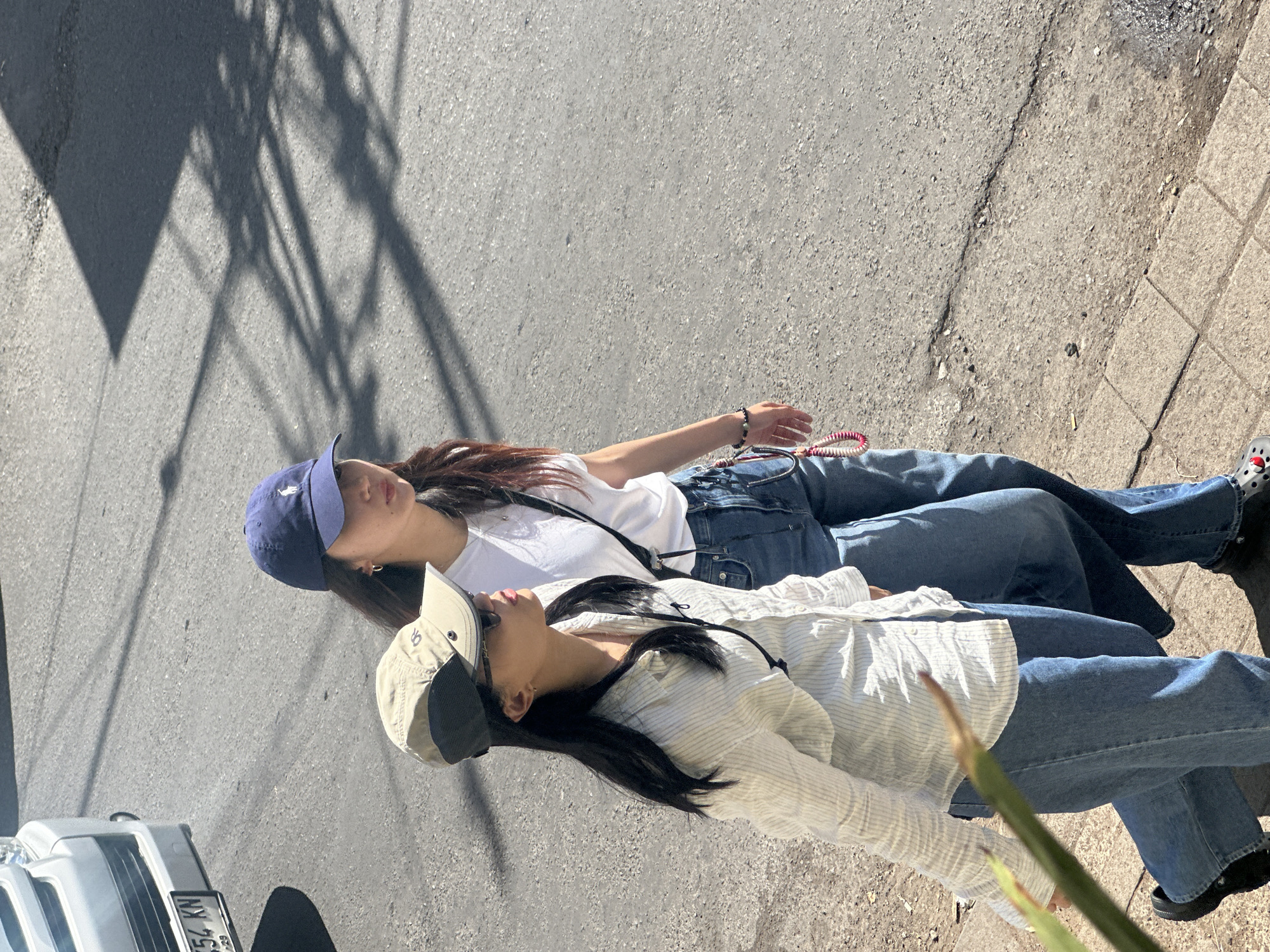 Two young women walk side-by-side on a paved street in what appears to be Ubud District, Indonesia, with the sun casting strong shadows. The woman on the left wears a light-colored striped button-down shirt, blue jeans, and a light-colored baseball cap with a dark brim. She has black hair and wears sunglasses. A black lanyard with a clip is visible around her neck. The woman on the right wears a white t-shirt, blue jeans, and a dark blue baseball cap with a white logo. She has dark hair and is wearing hoop earrings. A dark strap crosses her chest. She has a beaded bracelet with a pink and white charm hanging from her left hip. A white vehicle with a license plate reading "554 KN" is partially visible on the left side of the frame. The ground is asphalt with a concrete sidewalk on the lower left.