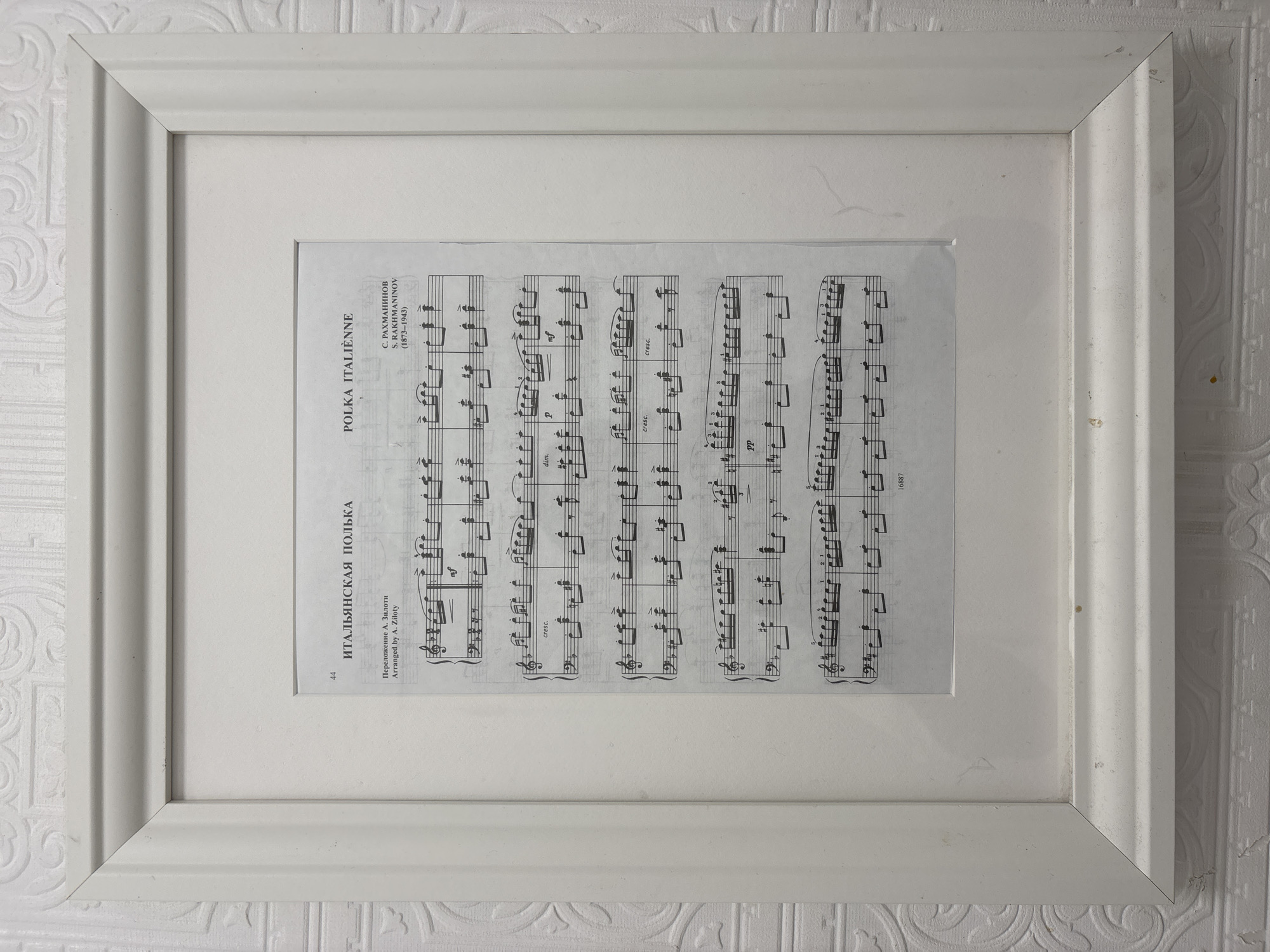 A framed piece of sheet music is mounted on a wall with patterned wallpaper. The frame is white and slightly weathered, with visible imperfections. The sheet music features the title "Polka Italienne" in both Russian and French, along with the name "S. Rachmaninov" and dates indicating the composer's lifespan (1873-1943). The music is arranged by A. Ziloty. The scene gives no clue as to the location.