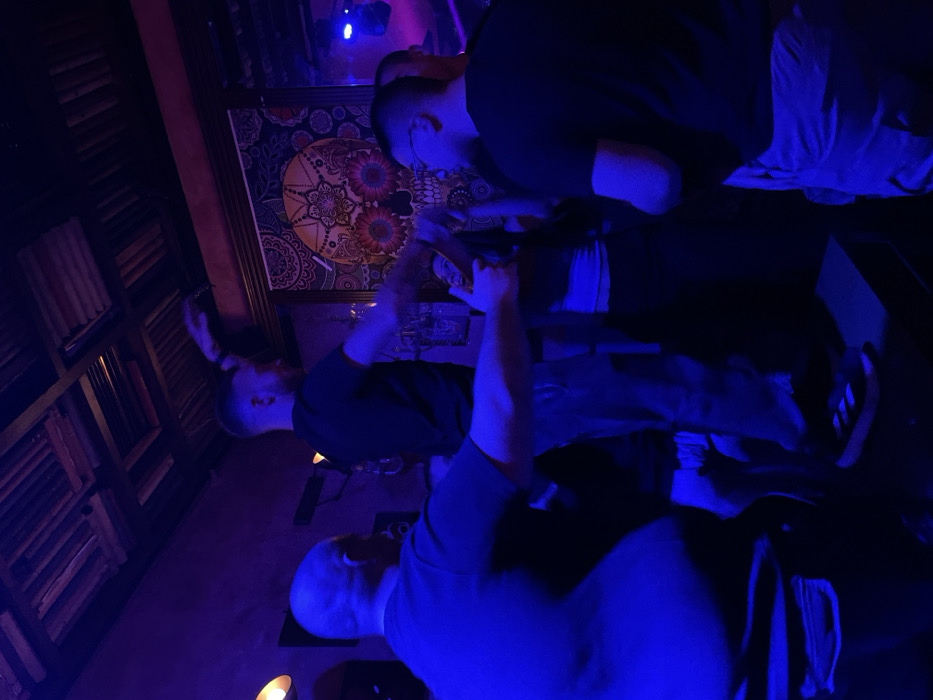 This image captures an indoor scene, likely a bar or lounge, at night in Drapetsona, Greece. The environment is dimly lit with prominent blue and purple artificial lighting, creating an atmospheric, club-like ambiance.
The primary subjects are several men, seen from various angles, engaged in a social gathering. In the lower right foreground, a bald man wearing a light-colored shirt is visible from behind and his side profile. To his left, another man with a shaved head, dressed in a dark shirt, stands with his left arm raised, possibly gesturing or holding an object over what appears to be a bar or high table. Further in the background, near the center, a third man in a dark shirt and glasses is seen from his back and side, leaning towards a surface where bottles or glasses are placed. A fourth figure, partially visible, is also present on the far right, with a foot wearing an Adidas sneaker being identifiable.
The setting is decorated with a large, framed piece of art on the right wall, featuring a colorful, intricate, mandala-like or floral design. On the left, there's a section of wall adorned with dark wooden paneling, possibly shelves or decorative louvers. A warm, glowing light fixture is visible low on the wall in the lower left quadrant. The activity appears to be casual socialization or enjoyment at a nightlife venue. There is no discernible text visible in the image.