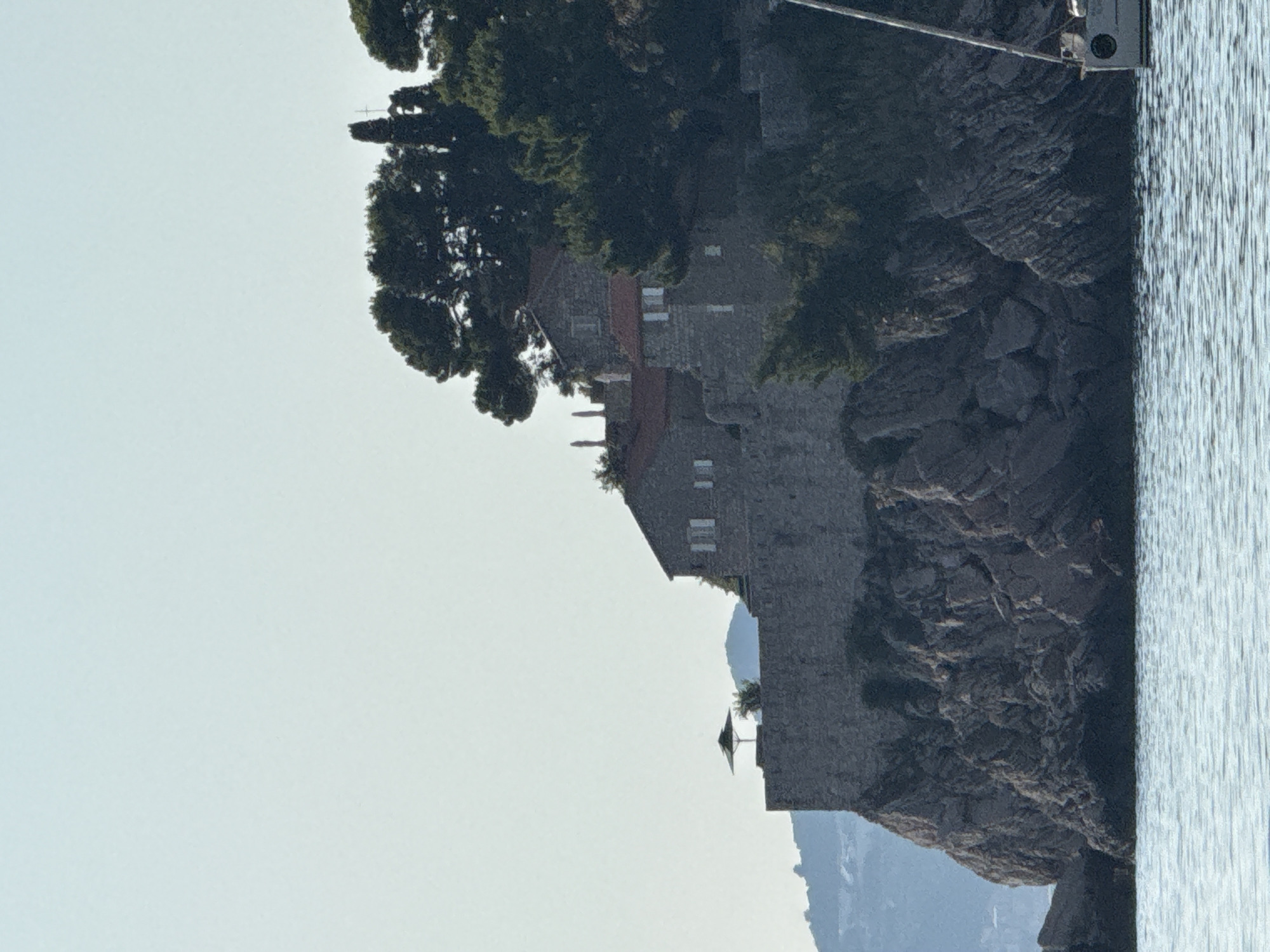The image shows a view of the island of Sveti Stefan, Montenegro, from the water. The island is a fortified village built on a rocky islet, connected to the mainland by a narrow causeway. In the foreground, the calm blue water of the Adriatic Sea reflects the clear sky. In the mid-ground, the rocky cliffs of the island rise steeply, topped with ancient stone buildings with red-tiled roofs. Lush green trees, including pine trees, grow on the slopes and around the buildings. A modern sailboat is partially visible on the right side of the image, its mast extending towards the top. In the distance, rolling hills covered in vegetation can be seen under a hazy sky. The overall scene depicts a serene and picturesque coastal landscape.
