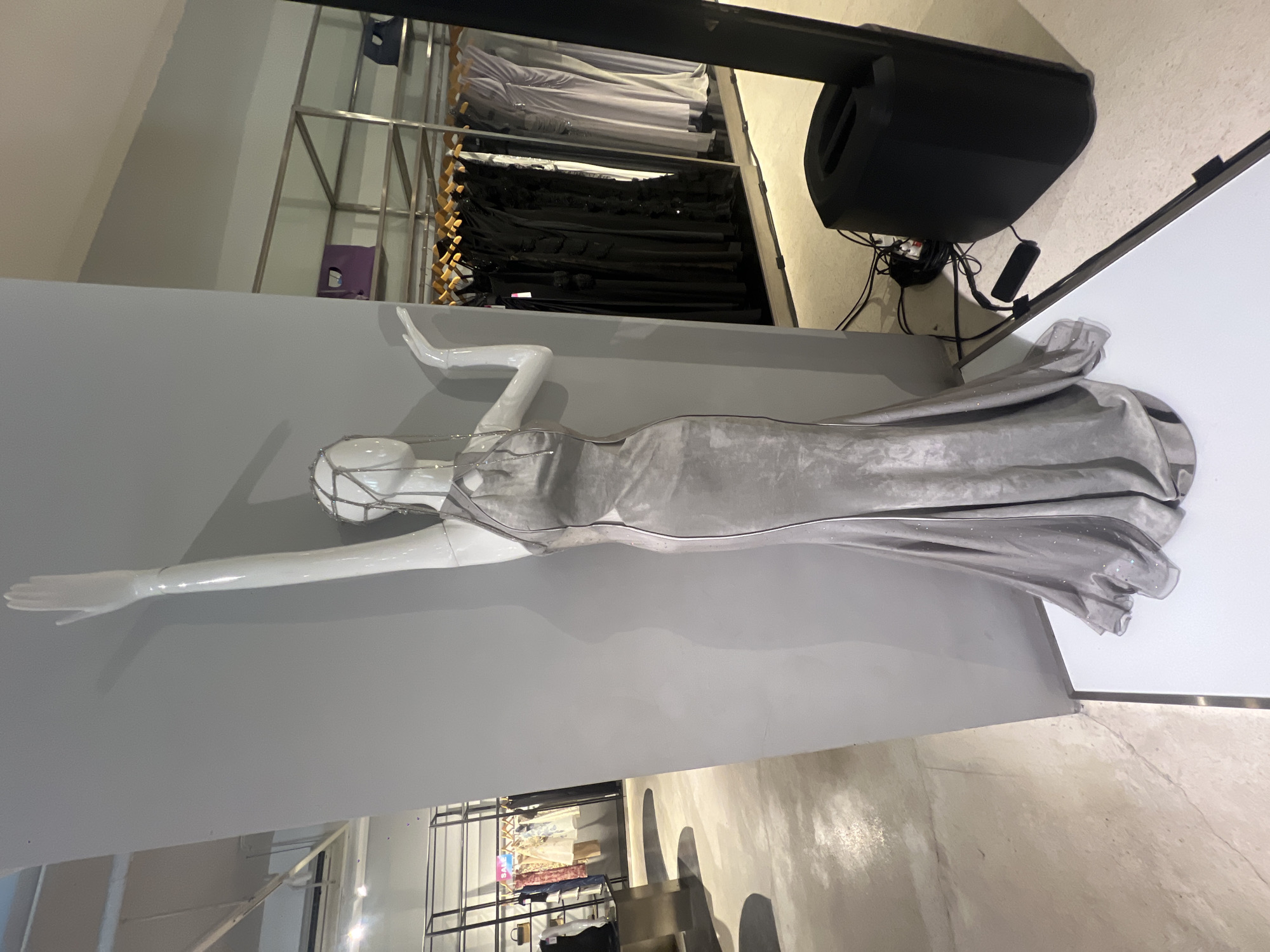 A mannequin in a silver gown reclines on a gray platform in a store. The store has clothing racks filled with various items. This could be a boutique in Kuta, Indonesia. A black speaker sits near the mannequin.