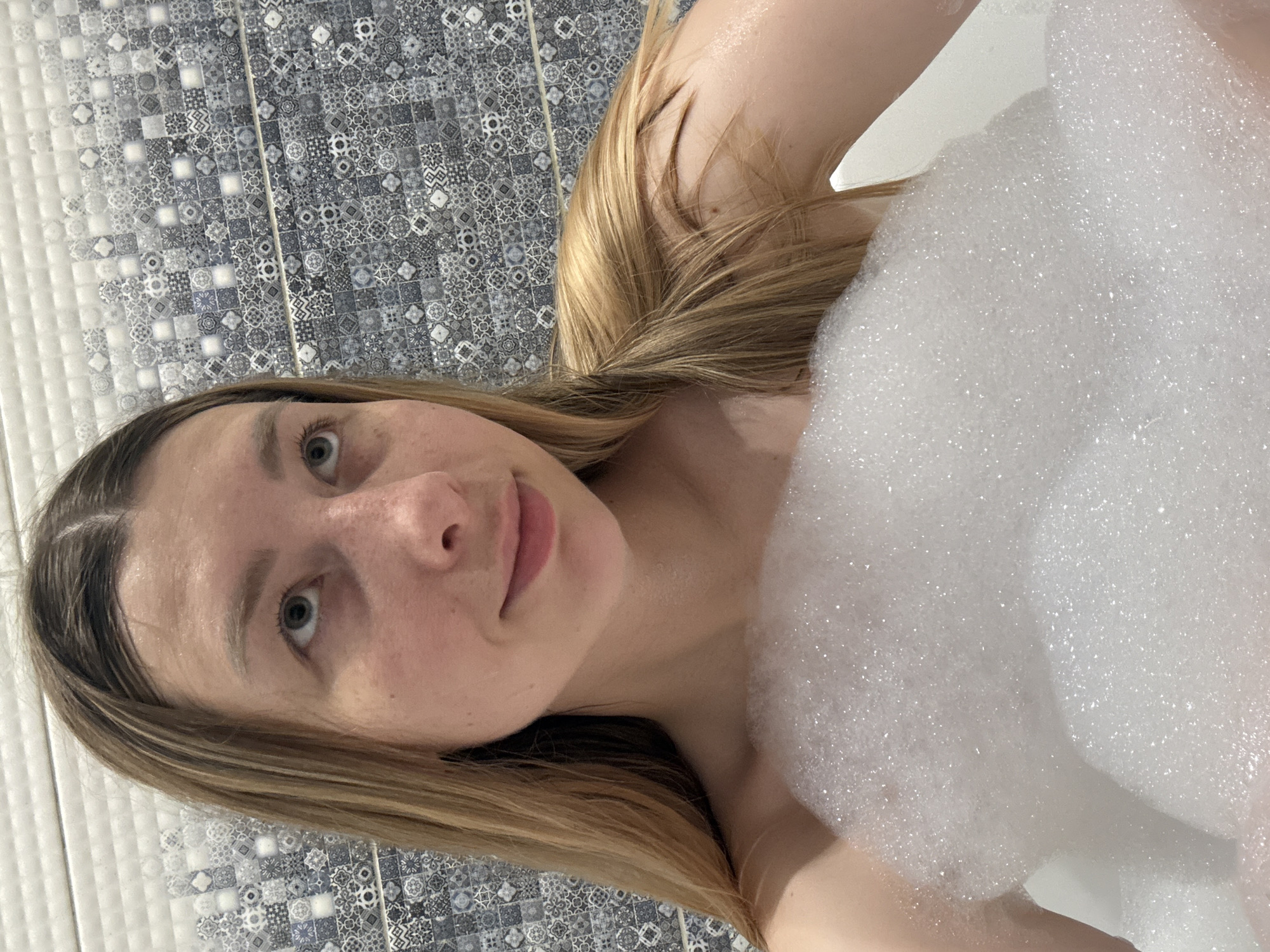 A woman is shown in a bathtub filled with bubbles. She has light-colored hair and is looking at the camera. The background consists of patterned blue and white tiles. There is no indication of location in the city of Moscow, Russia.