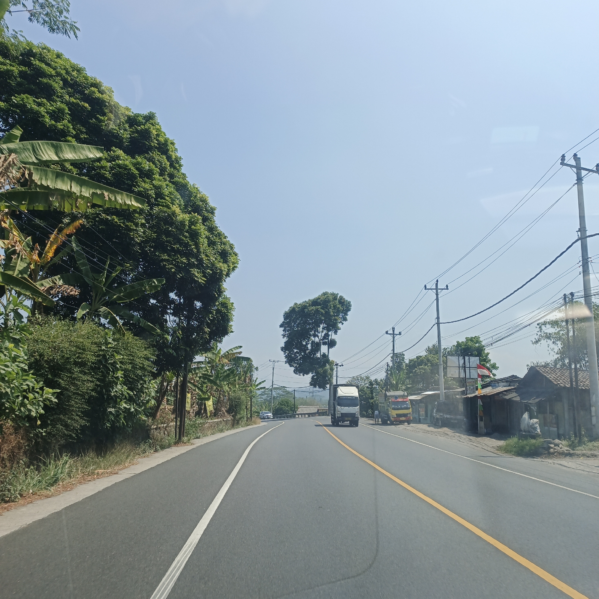 The image depicts a sunny day scene on a paved road in Pringsurat, Indonesia. The road has a white line on the left and a yellow line on the right, indicating traffic lanes. To the left of the road, lush green trees, including banana trees, and dense vegetation line the roadside. On the right side of the road, there are several small buildings with tiled roofs, power poles with numerous wires, and a few parked trucks, one white and one yellow. A small Indonesian flag is visible on one of the buildings. In the distance, more trees and hills can be seen under a clear blue sky. The perspective suggests the photo was taken from a vehicle moving along the road.