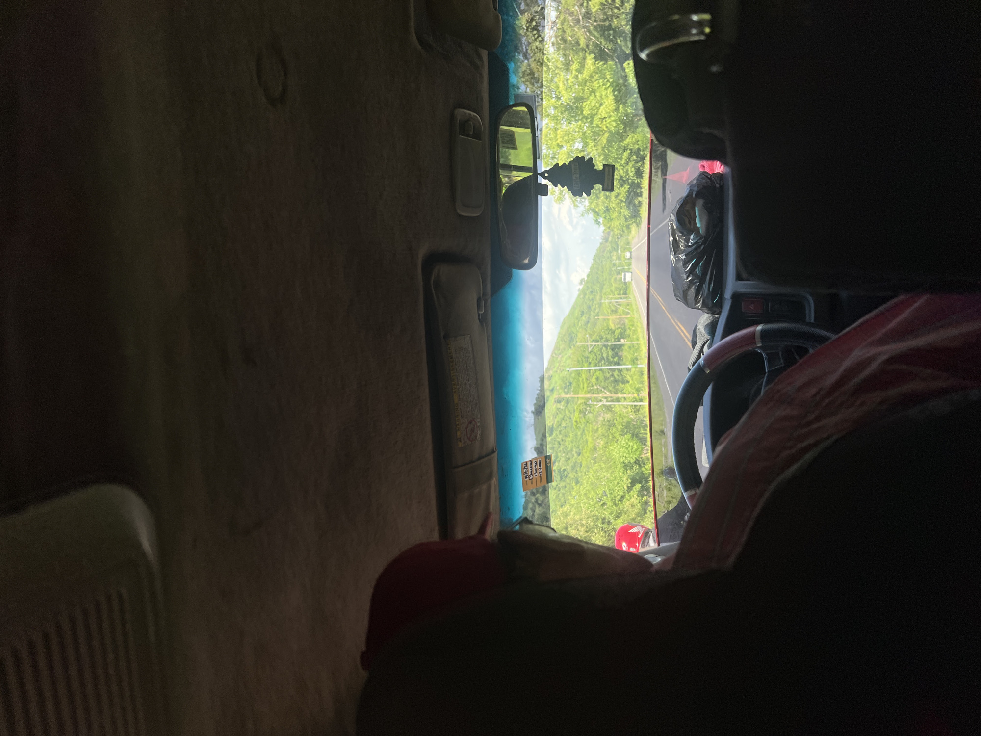 The image captures the interior of a vehicle, looking forward through the windshield at a road lined with lush green trees and rolling hills, indicative of a rural or semi-rural setting. The scene suggests a journey through San Lorenzo, Honduras, a region known for its tropical climate and scenic landscapes. The sky is partly cloudy, with a bright, sunny atmosphere.
In the foreground, a person wearing a red baseball cap and glasses is seated in the driver's seat, their back to the camera. They are wearing a pink and white striped shirt. Another individual, wearing a red helmet, is visible in the passenger seat. The dashboard of the vehicle is cluttered with various items, including a black plastic bag, a traffic cone, and a black air freshener hanging from the rearview mirror. A small yellow sticky note with writing on it is affixed to the windshield. The road ahead shows a few distant vehicles, suggesting moderate traffic. The overall impression is that of a casual road trip through a vibrant, green environment.