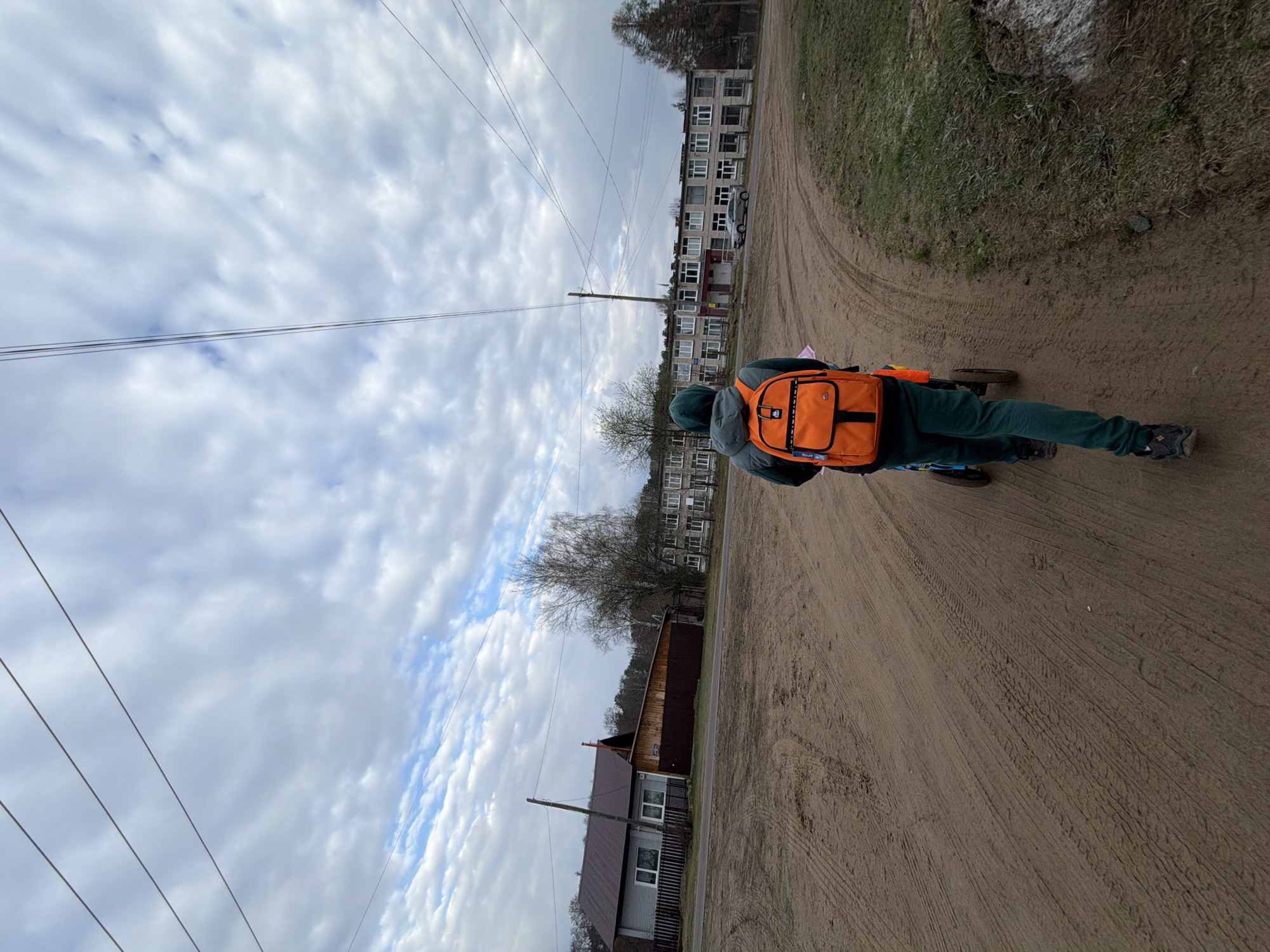 An outdoor, eye-level shot captures a person from behind, riding a low-slung, three or four-wheeled bike or scooter on a wide dirt road. The person is dressed in a dark green hooded top and matching pants, with dark sneakers. They are carrying a vibrant orange backpack, which features a prominent white tag displaying the text "ROLLIN' ON" and possibly "ARMS" or "APMS" beneath it. The wheeled vehicle has a blue frame, and its rear wheels are visible below the rider.
The unpaved road, marked with numerous tire tracks, stretches into the distance. On the left side of the road in the mid-ground, a single-story house with a grey roof and light-colored walls, partially obscured by fencing and bare trees, can be seen. Further in the background, lining the right side of the road, is a long, multi-story apartment building with many windows, indicative of Soviet-era architecture commonly found in locations like Chaikovsky, Russia. A white car is parked in front of this building. Utility poles and multiple electrical wires crisscross the expanse of the sky.
The sky is predominantly overcast with soft white clouds, yet patches of blue are visible, suggesting a bright, partly cloudy daytime. The trees in the scene appear mostly bare, hinting at either early spring or late autumn. The overall setting depicts a tranquil, semi-urban or suburban environment.