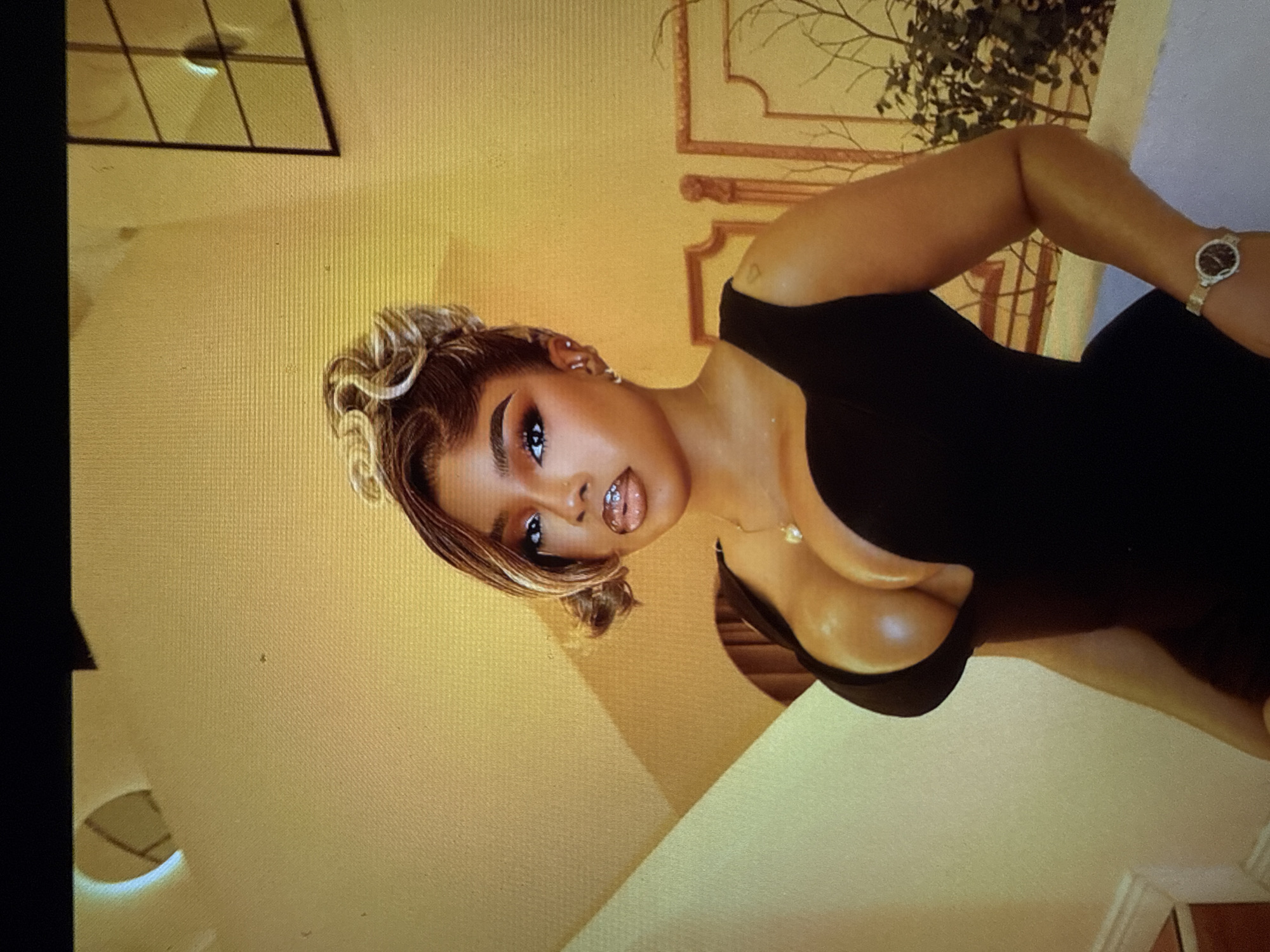 A woman is shown in a medium shot, facing the camera. She is wearing a black, low-cut dress and has her hair styled in an updo with loose curls framing her face. Her makeup is dramatic, with bold eyeshadow and a glossy lip. She is also wearing earrings and a necklace with a heart-shaped pendant. A watch is visible on her left wrist. The background appears to be a staircase in an indoor setting, with a wall painted a warm yellow color. There is a framed picture on the wall behind her. The lighting is warm and highlights the woman's features and the texture of the wall. The image quality suggests it may be a photograph taken from a screen, as evidenced by the pixelation and slight glare.