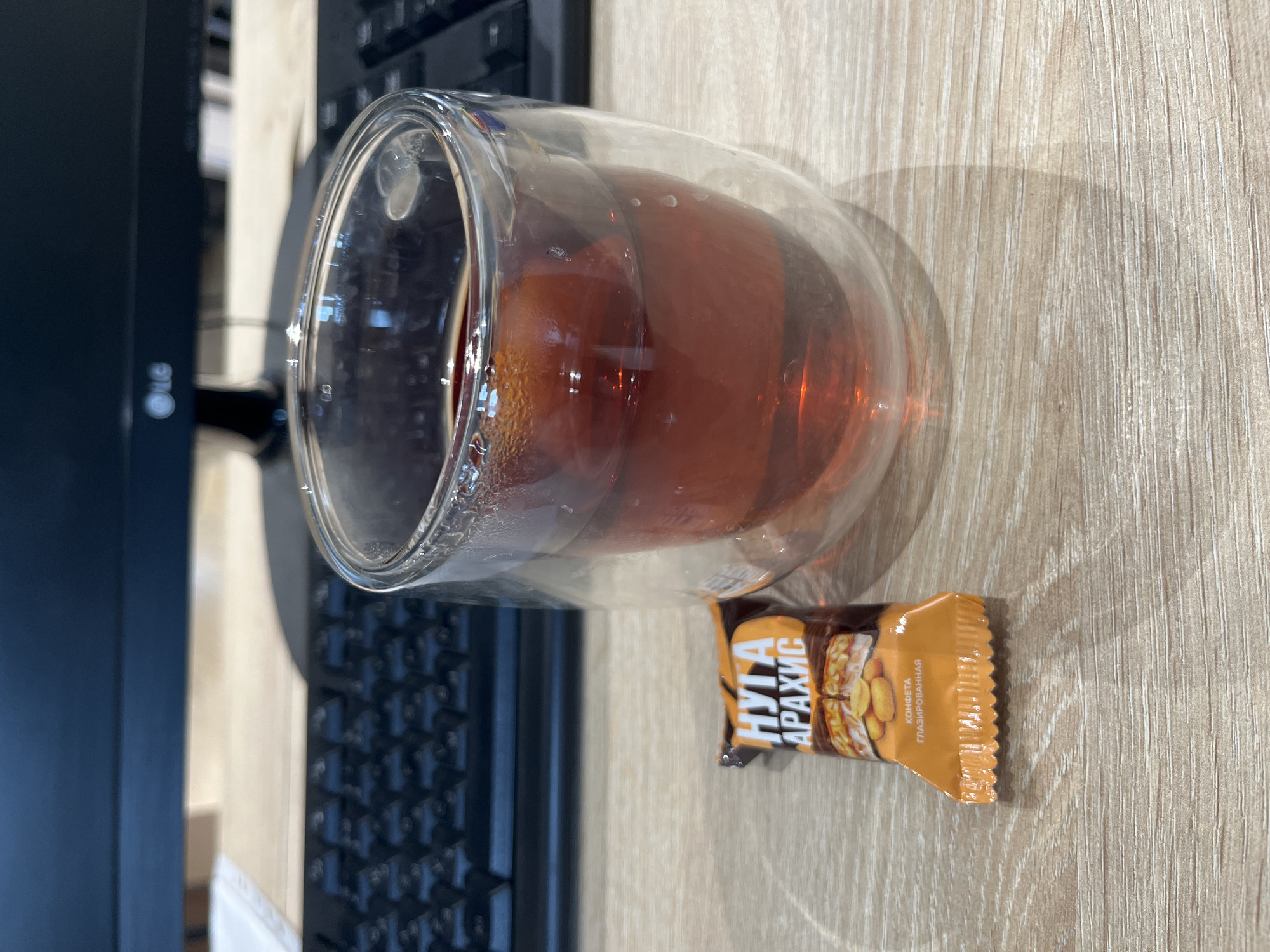 The image shows a desk with a glass cup filled with a dark liquid, possibly tea. Next to the cup is a candy bar with a tan wrapper and text written in a language other than English. A black keyboard is partially visible, and a black computer monitor is present in the background on the left side of the image. The desk has a light wood grain pattern.