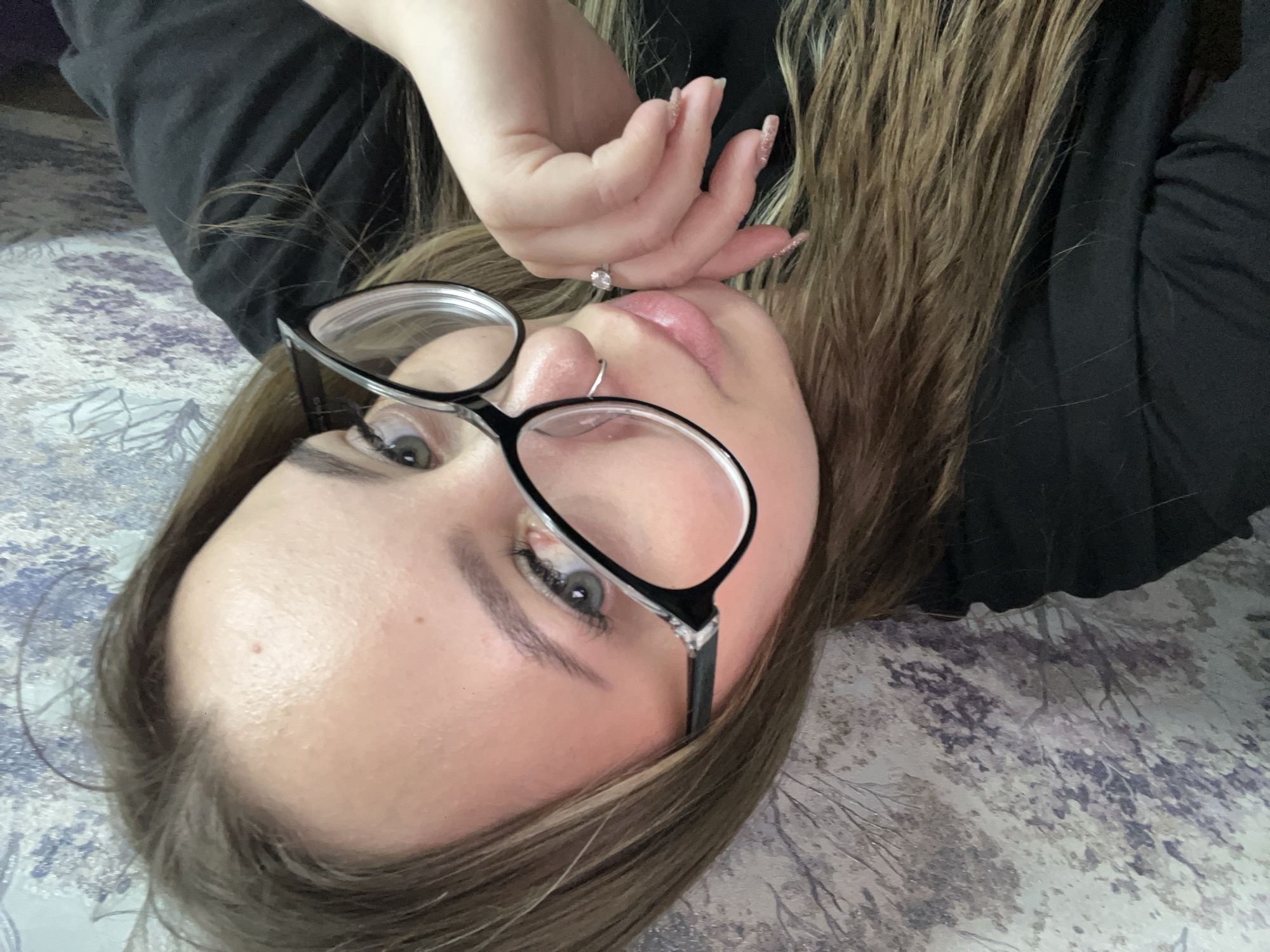 A fair-skinned female with blond/brown hair is seen lying on what appears to be a patterned bedspread. She is wearing glasses and a black shirt. Her left hand is near her mouth. A nose ring is visible. A ring is visible on her hand. The image does not contain information to confirm it was taken in Novopokrovskaya, Russia.