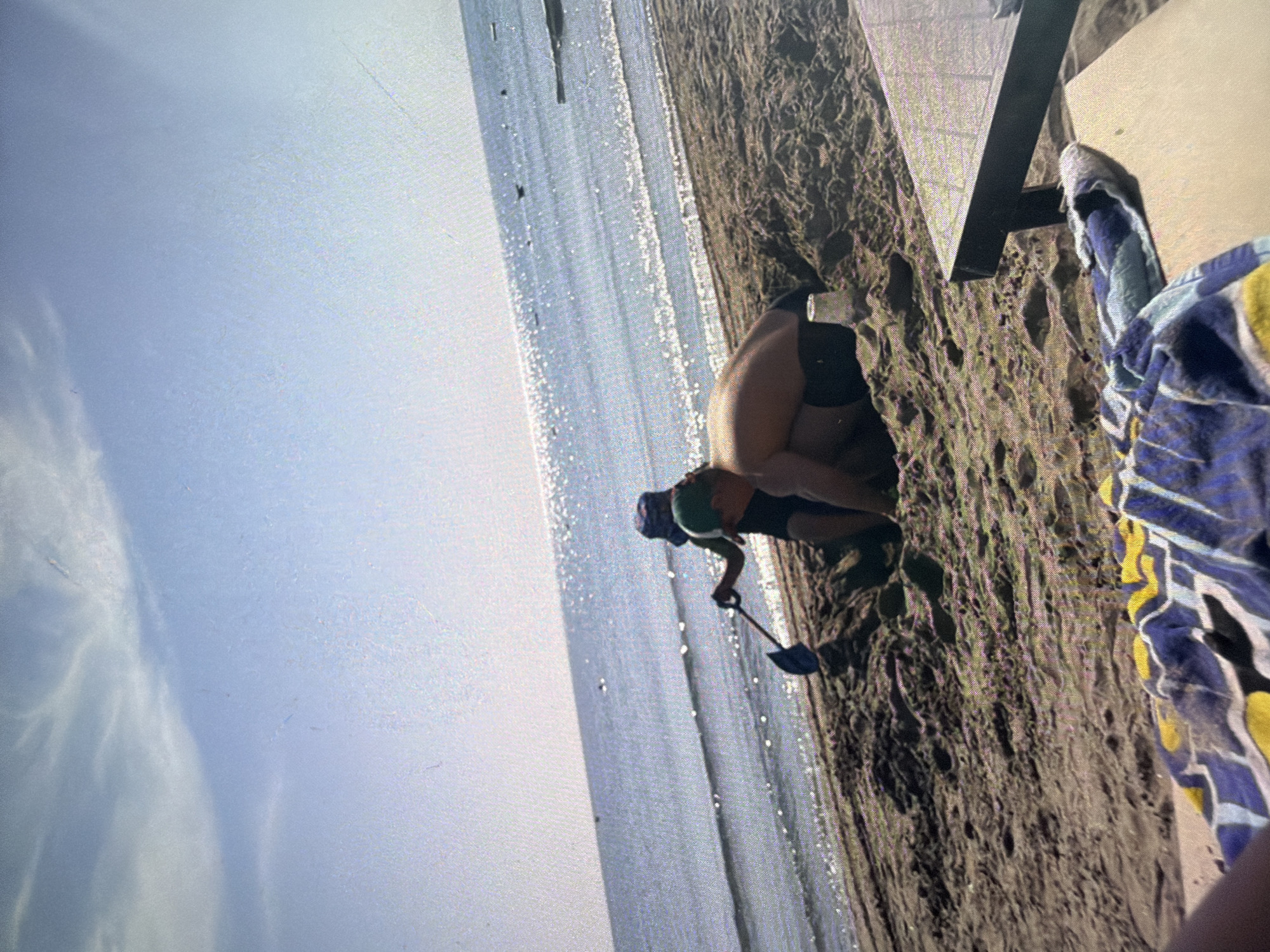 Two individuals are on a sandy beach, with the ocean and sky visible in the background. One person, wearing a blue and white patterned hat, is digging in the sand with a blue shovel. The other person, wearing a green hat and black shorts, is lying on their stomach and digging in the sand. A blue and yellow patterned towel is in the foreground. A dark-colored lounge chair is partially visible on the right side of the image. The scene is under a bright, partly cloudy sky.