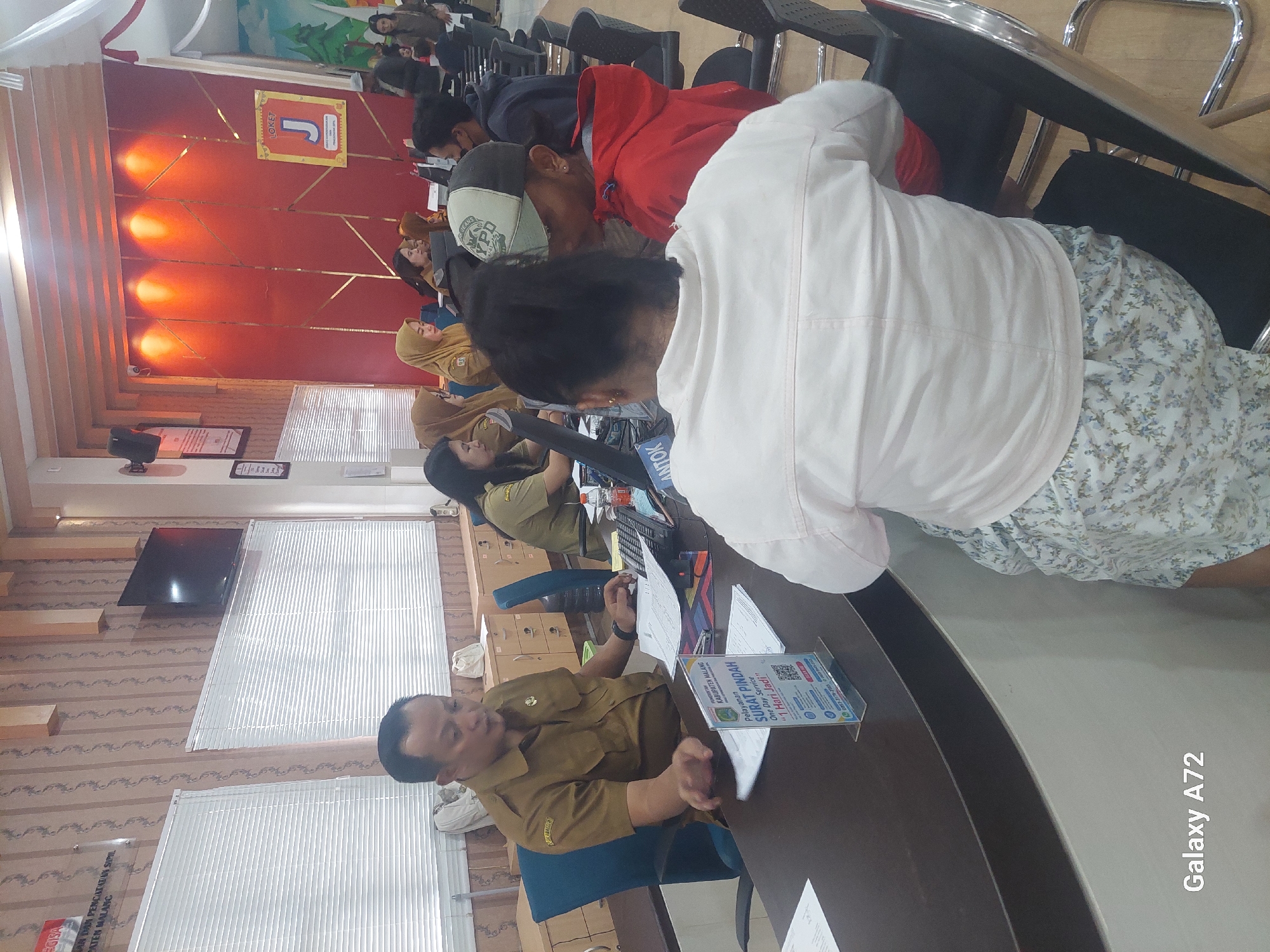 In the city of Kedungpedaringan, Indonesia, within a government office, a male official wearing a khaki uniform sits at a desk and appears to be interacting with a woman seated opposite him. The official is gesturing with his right hand while looking at documents. In front of him, a clear plastic stand displays information, including text in Indonesian and a QR code, with the words "Pelayanan SURAT PINDAH" prominently visible. The desk also holds a calculator and papers. In the background, other individuals are seated at desks, seemingly engaged in similar administrative tasks, with several women wearing headscarves. The office environment features blinds on windows and what appears to be a television mounted on the wall. The overall scene depicts a busy public service counter.