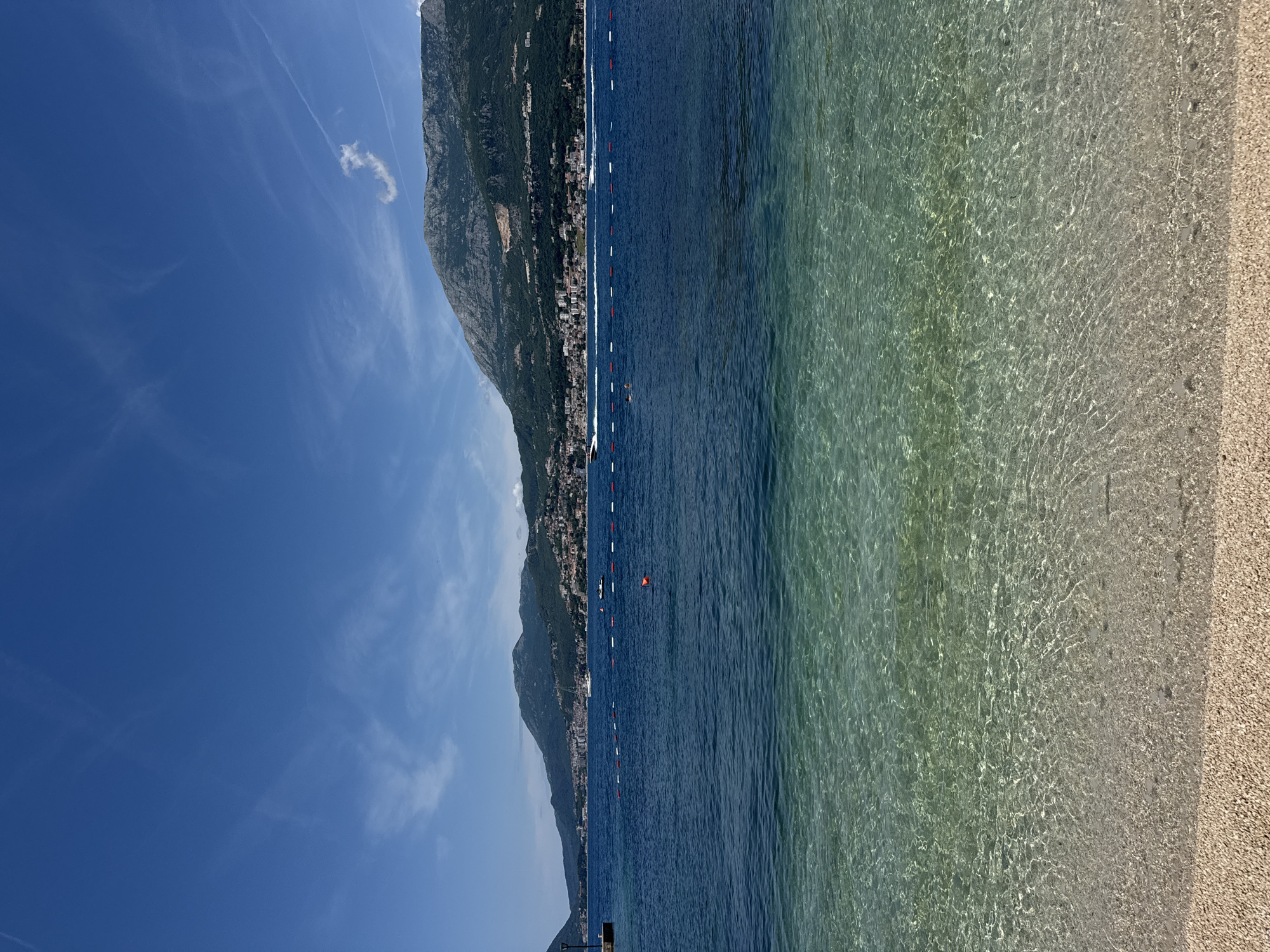 The image displays a bright, sunny coastal scene featuring a pebble beach, clear water, a bay with boats, a distant town, and mountainous terrain under a blue sky.
In the foreground, a light-colored pebble beach transitions into exceptionally clear, shallow water that reveals the rippled seabed beneath. The water gradually deepens, becoming a darker blue-green. A horizontal line of red and white cylindrical buoys marks a boundary in the mid-water, with a single orange spherical buoy positioned to its right. Two small, dark figures, consistent with swimmers, are present near the buoy line.
Several boats are visible on the water: a white speedboat moves from left to right, creating a prominent white wake, while a smaller white boat is further to the right. A white sailboat is either anchored or drifting slowly in the distance on the left side of the bay. On the far left, a dark post stands on a small jetty or pier.
The background features a continuous stretch of land. A town or city with numerous buildings is built along the coastline and extends up the lower slopes of the adjacent mountains. These mountains are rugged, predominantly covered in green vegetation, and show areas of exposed rock. The sky is a clear, vibrant blue with scattered wispy white clouds and several thin, linear contrails. The overall impression is of a calm, warm, and clear day at a coastal location. The specific city and region remain unknown from the image content.