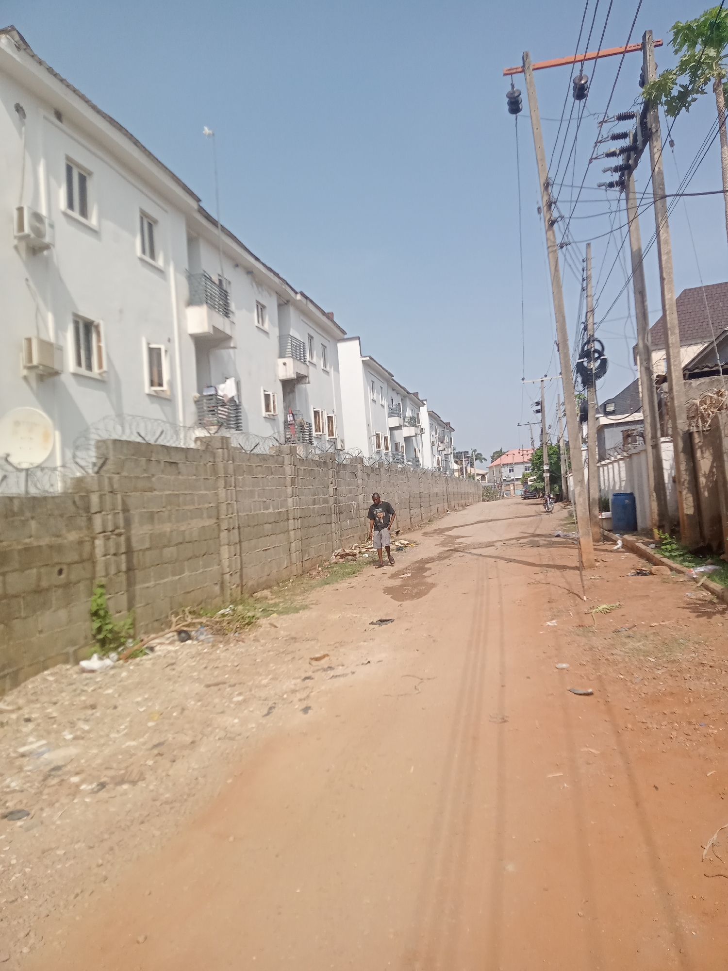 The image captures a clear, sunny daytime scene of an unpaved, reddish-brown dirt road in a residential area, identified as Gwarinpa, Nigeria. The road, showing distinct tire tracks, stretches into the distance, bordered by structures and natural elements.
On the left side of the road, a man wearing a dark t-shirt with an indiscernible graphic and light shorts walks away from the camera, seemingly carrying a small bag or object. He is positioned near a robust concrete block wall, topped with extensive barbed wire, which acts as a boundary for a row of multi-story white residential buildings. These buildings feature multiple windows, balconies with metal railings, and external air conditioning units. A white satellite dish is also visible near the wall on the left.
The right side of the road is lined with several tall wooden utility poles, heavily laden with a complex network of electrical wires and insulators. Scattered debris and a blue plastic barrel are visible on the ground near these poles. Further down the road, additional residential buildings with varied architecture and roof colors can be seen. The sky above is clear and light blue, indicating fair weather. The scene suggests a bustling residential neighborhood with developing infrastructure. No explicit text is clearly visible on any signs or objects within the frame.