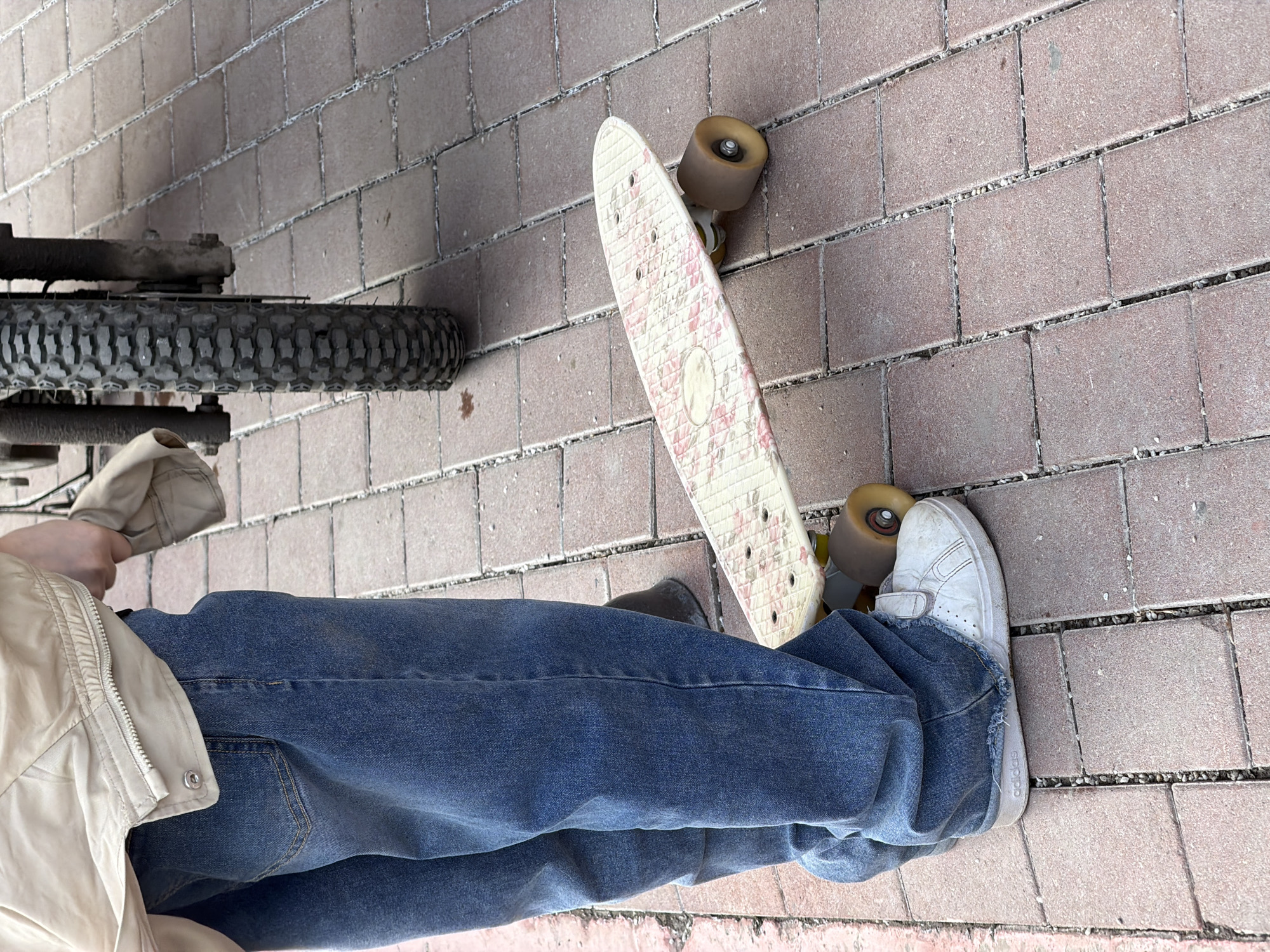 The scene is a close-up, tilted overhead shot of a person standing on a brick-paved surface. The person is wearing blue jeans, a beige jacket, and white sneakers. A white skateboard with a floral pattern is next to their foot. A bicycle wheel is partially visible on the left side of the frame. The location is possibly Moscow, Russia, suggested by the type of brick paving, though not explicitly confirmed. The environment suggests an urban setting.