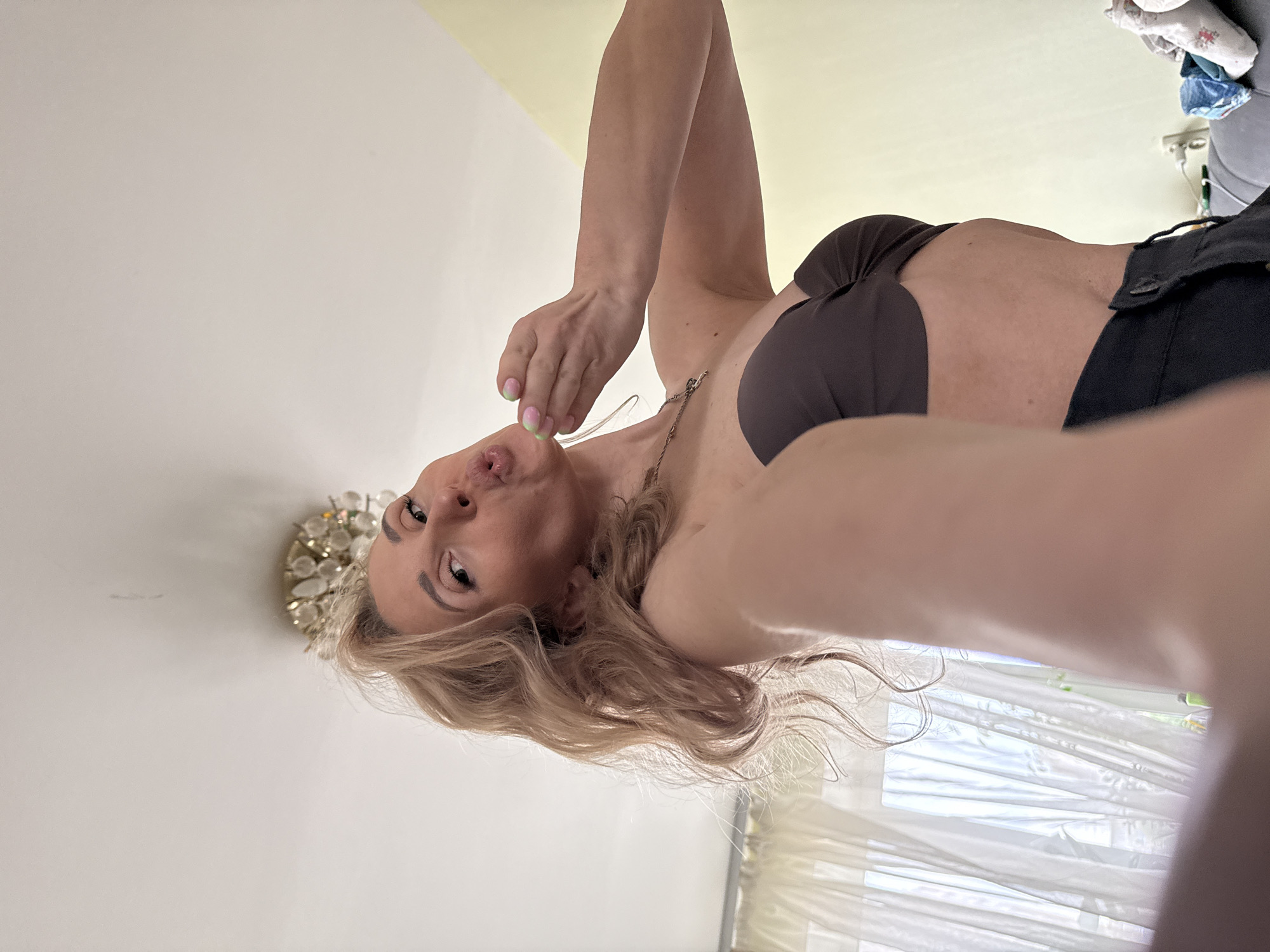This media file features a selfie of a woman taken indoors, likely in Chaikovsky, Russia. The woman, who has blonde or light brown wavy hair and a light skin tone, is captured from a low-angle perspective, giving the impression that she is looking down at the camera. She is wearing a dark brown or black strapless top or bra and dark pants. A thin silver necklace is visible around her neck. Her fingernails are painted with a distinct gradient of pink and light green. She is pouting her lips, as if blowing a kiss or making a playful expression, with one hand raised near her mouth.
The background primarily consists of light-colored walls, possibly white or light beige. Behind the woman's head, a decorative wall-mounted light fixture with multiple shimmering elements is visible. To the right, a window with sheer white curtains diffuses natural light into the room, suggesting it is daytime. In the upper right corner, some items, possibly folded clothing, are stacked on a surface. The overall setting appears to be a casual domestic environment, such as a bedroom or living room. The image conveys a playful and relaxed mood. There is no visible text in the media file.