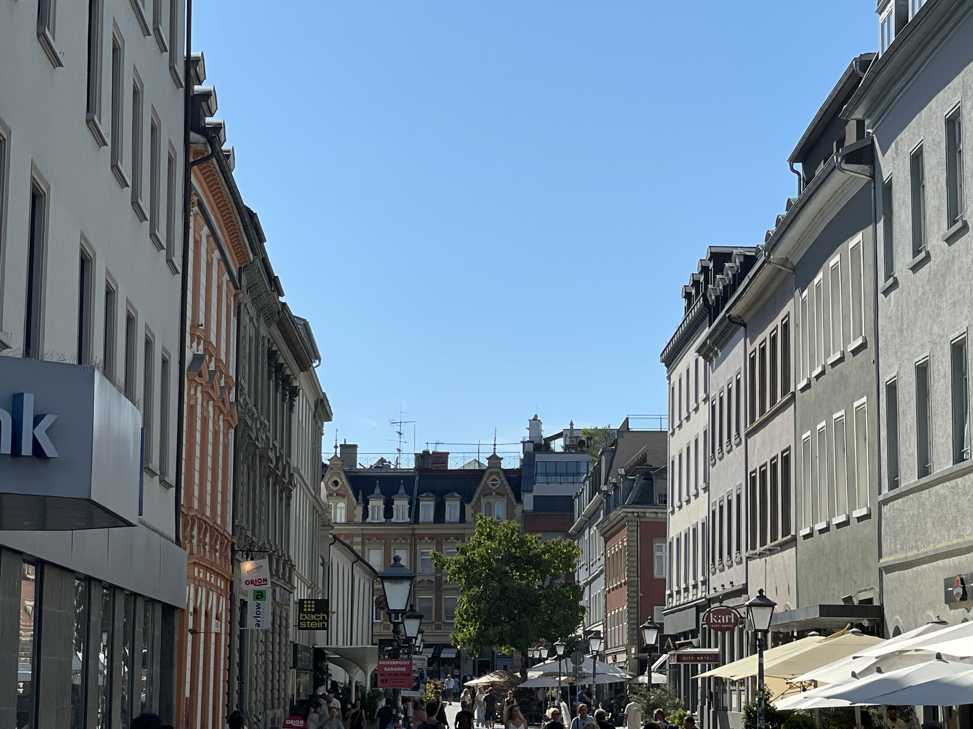 The image depicts a street scene in the city of Constance, Germany. The perspective is from a street looking forward, with buildings lining both sides. On the left, a modern building with a prominent "bank" sign is visible, adjacent to older buildings with orange and grey facades. The right side of the street is dominated by a row of older, multi-story buildings with grey and pastel-colored facades. Several shops and businesses are indicated by signs, including "Orion," "Bachstein," and "Karl City-Hotel." In the background, more buildings with distinct architectural features, including dormer windows and mansard roofs, are visible. A lush green tree stands in the middle ground, surrounded by outdoor seating areas with umbrellas. People are walking along the street, some appearing to be tourists, and others patrons of the establishments. The sky is clear blue, indicating a sunny day. The overall impression is that of a bustling European city street on a pleasant day.