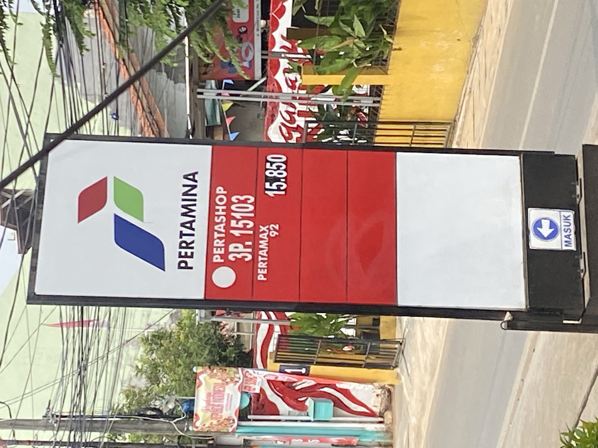 An eye-level medium outdoor shot captures a red and white Pertamina signpost in Tangerang, Indonesia. The sign displays fuel prices and the Pertamina logo. To the left of the sign, a narrow street runs parallel to the signpost, with a yellow wall and a black metal fence in the background. Several trees are visible behind the fence. A few power lines are strung across the top of the frame, obscuring a light green building in the background. The sky is overcast.