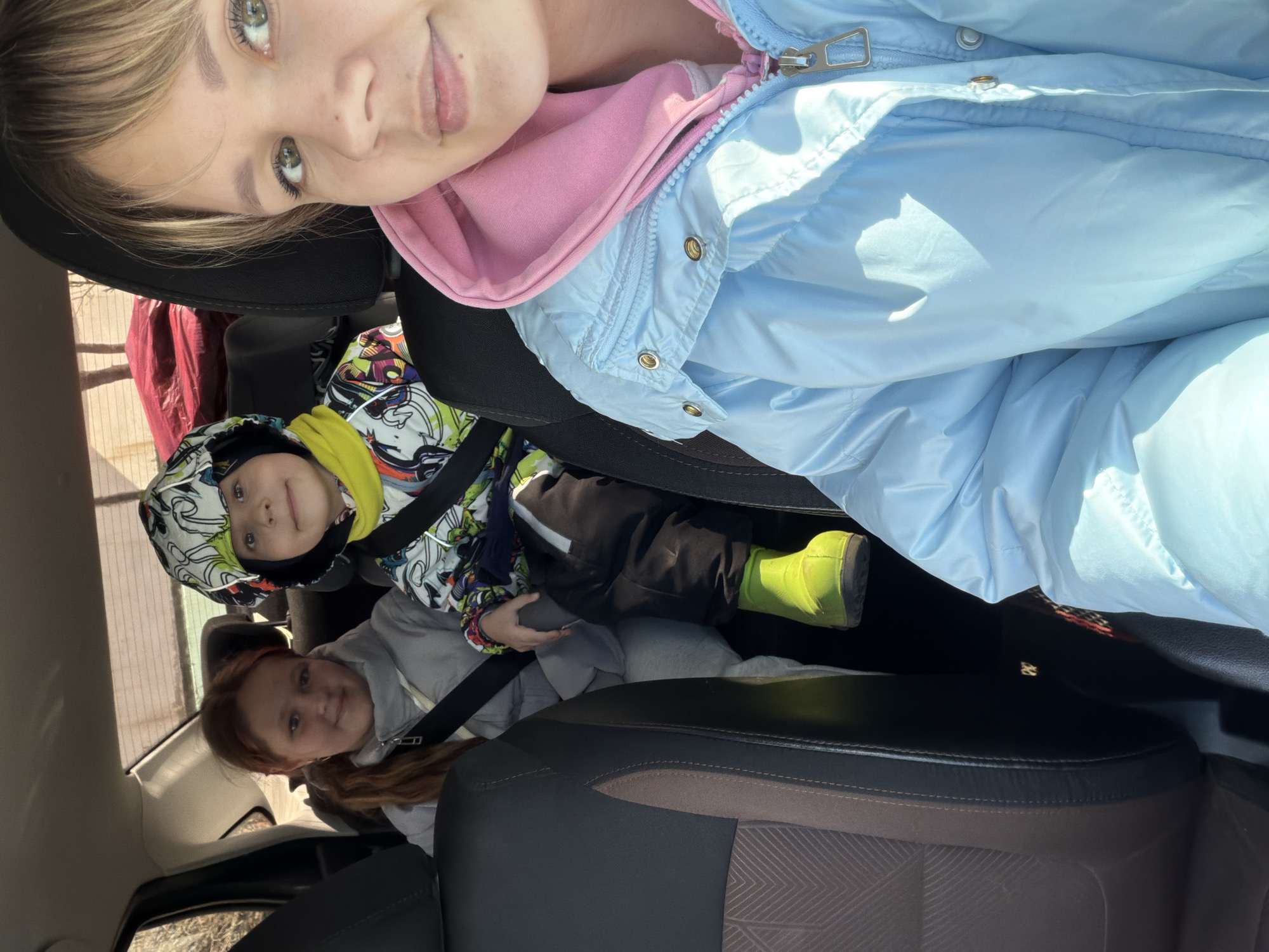 A mother and two children are in a car. The mother, wearing a blue jacket over a pink hooded sweatshirt, is in the front seat. The children are in the back seat; one is wearing a patterned hat and a yellow jacket, while the other wears a gray jacket. All three are facing the camera. All are wearing seatbelts. The location is unknown.