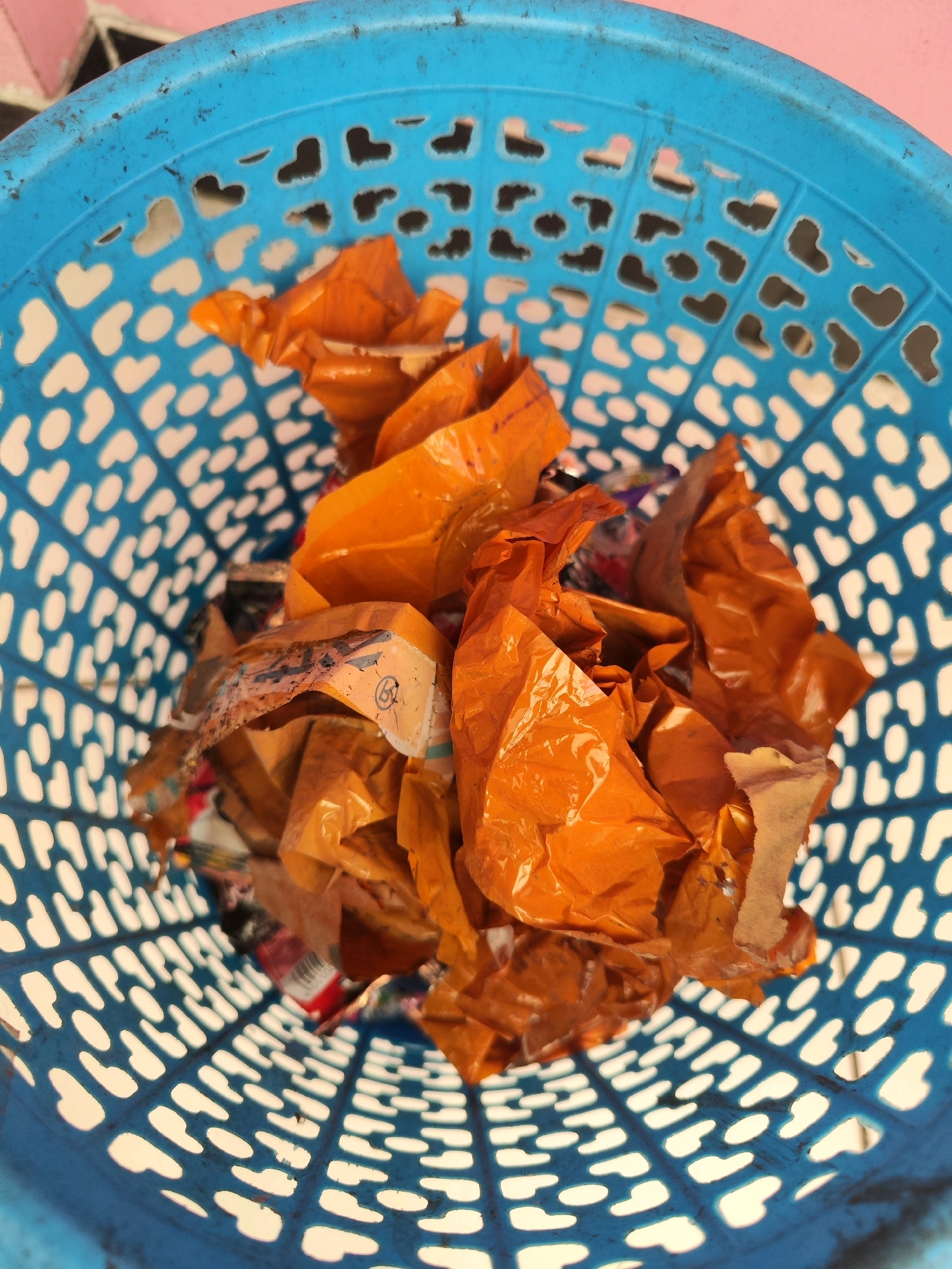 A close-up, overhead shot shows the interior of a blue plastic trash can filled with crumpled orange plastic wrappers and other miscellaneous debris. The trash can has a patterned surface with heart-shaped cutouts. The scene appears to be outdoors, possibly in Sragen, Indonesia, with a blurred background suggesting a pinkish wall and a tiled surface. There are no people or animals visible in the image.