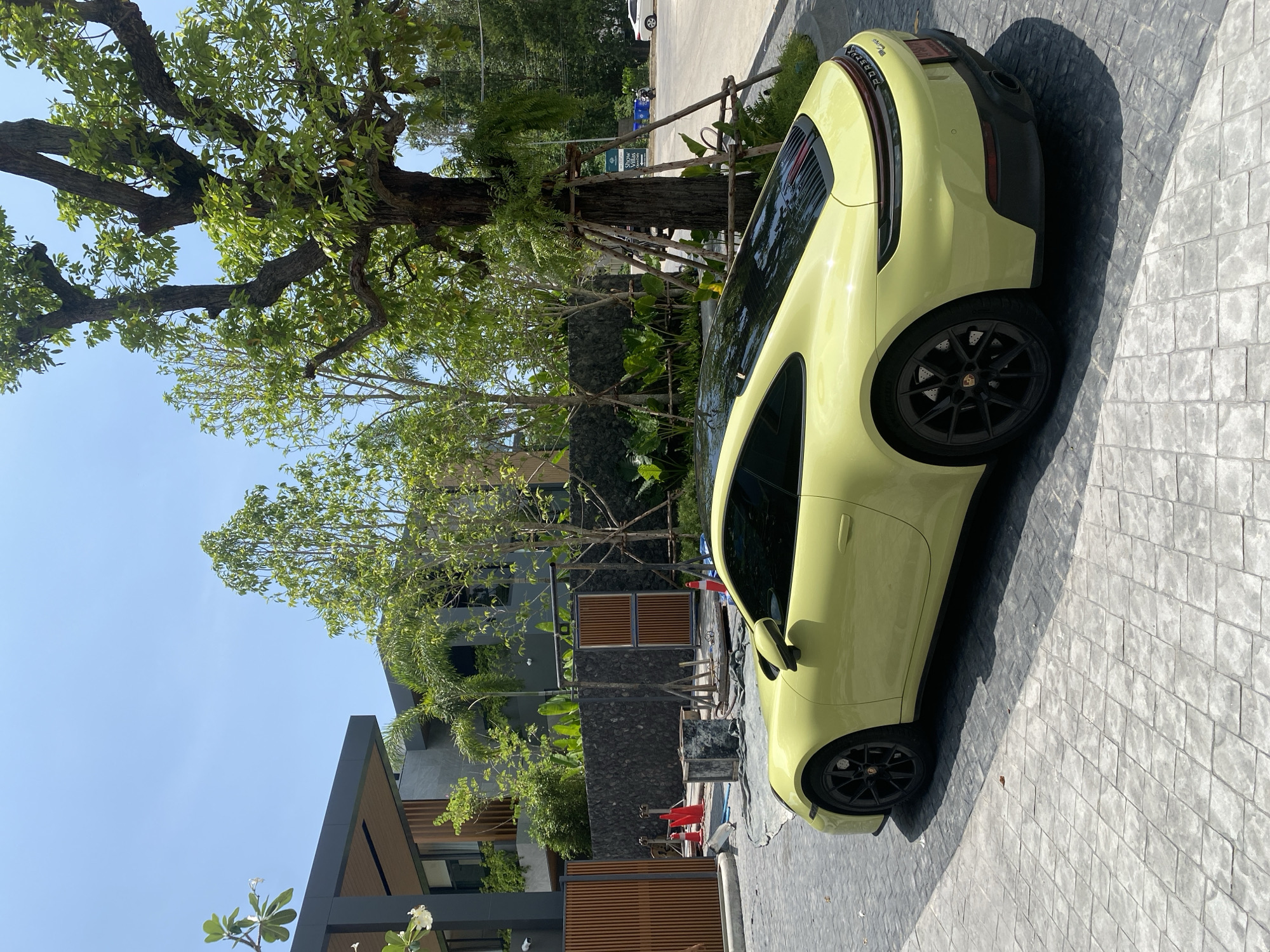 This vertical image, set in Choeng Thale, Thailand, features a light yellow (or pale green) Porsche 911 sports car parked on a patterned gray paving stone driveway. The car is positioned with its rear and right side prominently displayed, casting a long, sharp shadow on the ground, indicative of bright midday or early afternoon sun under a clear blue sky.
In the background, a lush and verdant environment is dominated by large, mature trees with dense green foliage, alongside smaller trees and palm fronds. Behind the greenery, modern residential buildings with dark gray facades, wooden slat accents, and stacked stone walls are visible, suggesting a contemporary and upscale architectural style. Construction or maintenance materials, including orange cones, buckets, and a tarp, are present near the car, hinting at ongoing work on the property.
Further in the distance, a white car and a blue scooter can be seen on a road or extended paved area. A small sign in the background, visible in the hint crops, identifies the location as "SHREW MANSION" and "VILLAS," confirming it as a residential complex. The Porsche itself features black multi-spoke wheels with visible brake discs and calipers, and "PORSCHE" badging on its rear, along with a blurry Thai license plate. No people are visible in the scene.