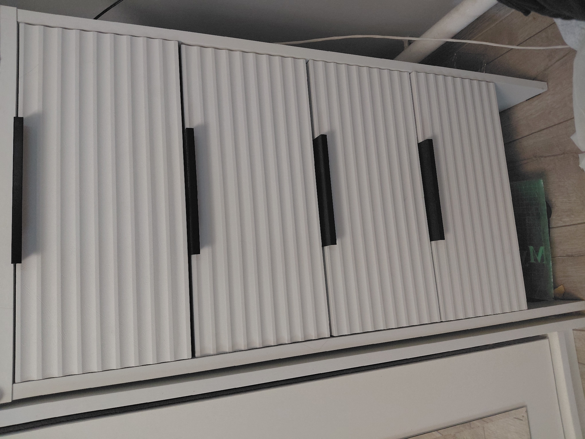 The image shows a white dresser with horizontal ribbed drawers and black handles. The dresser is positioned indoors, on a light wooden floor. To the left of the dresser, a portion of a white wardrobe or cabinet is visible. A grey wall and a white pipe are present in the background. The scene is static and appears to be a bedroom or storage area. There are no people or animals present in the image. The location is the city of Malinovka, Russia.