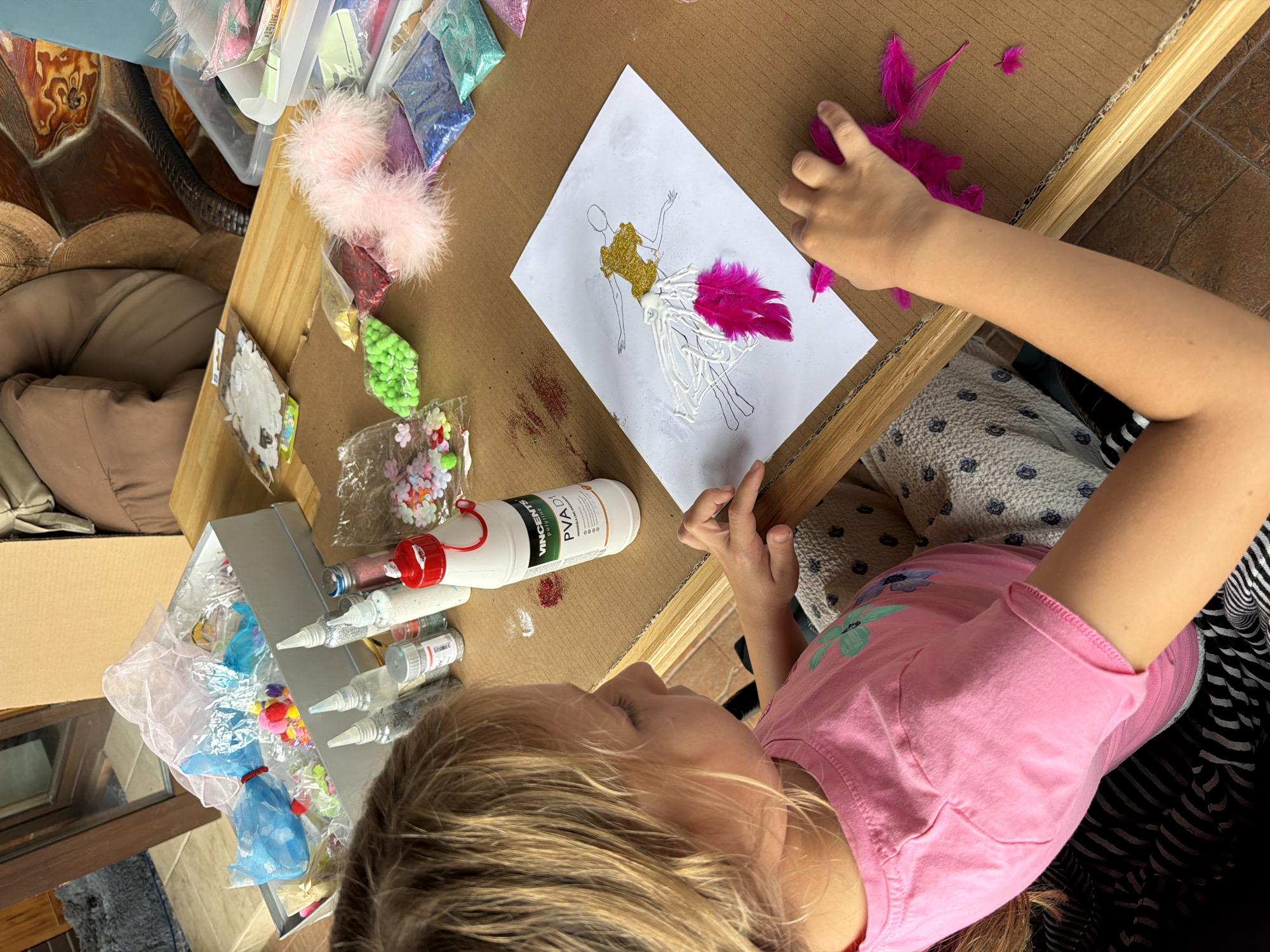 A young girl with blonde hair is sitting at a wooden table and crafting. She is wearing a pink t-shirt. The table is covered with various craft supplies, including glitter, feathers, small flowers, glue, and other decorative items. The girl is holding a pink feather and appears to be in the process of decorating a drawing of a figure wearing a dress. The background shows a blurry indoor setting with some furniture and boxes. This scene is located in the city of Baloži, Latvia.