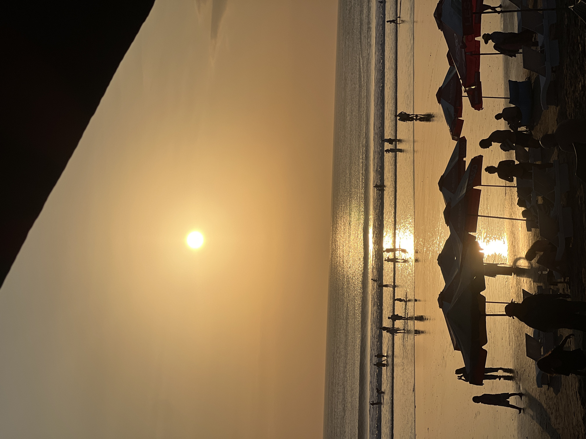 The image depicts a sunset scene at Kuta Beach in Kuta, Indonesia. The sun is a bright, round orb low on the horizon, casting a golden glow across the sky and reflecting on the ocean surface. The ocean is relatively calm with gentle waves rolling onto the sandy shore. Several people are visible in the water, wading and standing, their silhouettes stark against the bright water.
On the beach, more figures are scattered, some walking, others appearing to be interacting. In the foreground, a line of beach umbrellas, some red and white, are set up, providing shade over empty lounge chairs. The beach itself is wet near the water's edge, with puddles reflecting the sunset. The overall atmosphere is serene and evokes the end of a day at the beach. The image is captured from a slightly elevated perspective, looking out towards the ocean.