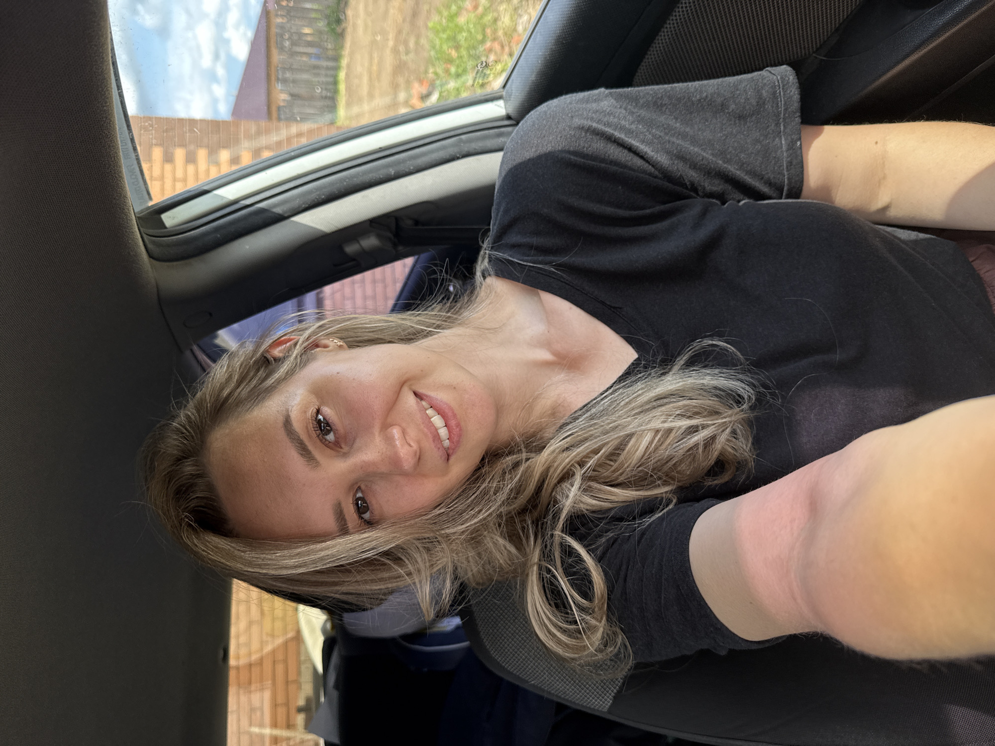 A woman with long, blonde, wavy hair is sitting in a car. She is wearing a black t-shirt and smiling. The car is parked near a brick building and a wooden fence in Chaikovsky, Russia. The sky is partially visible through the car window. The woman appears to be taking a selfie. Her left arm is visible and looks somewhat sunburned.