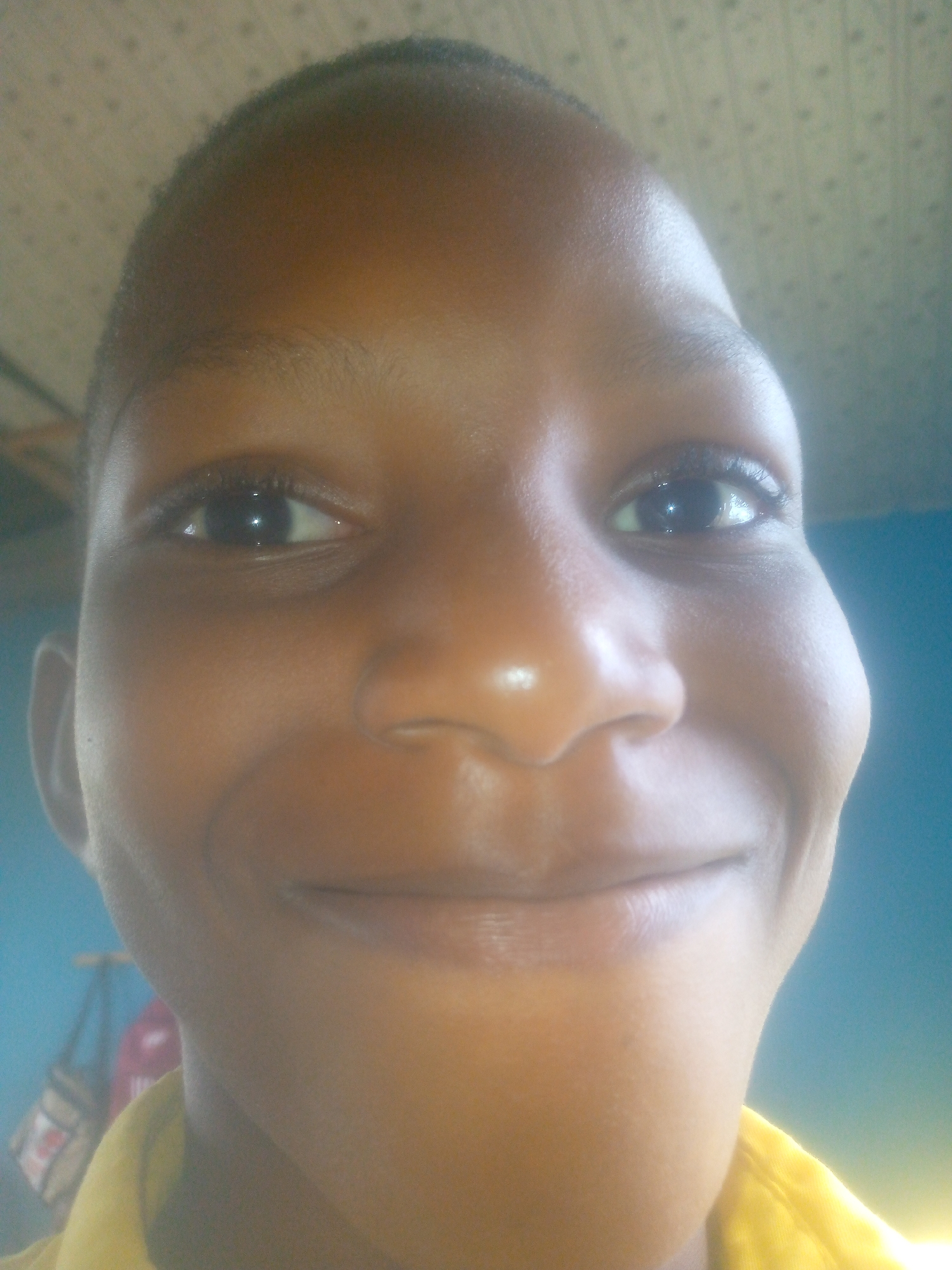 A close-up portrait of a smiling Black person with a shaved head. The background appears to be the interior of a room with a textured ceiling and blue wall. A yellow garment is visible around the person's neck and shoulder. A blurry bag hangs behind the person to the left. The location is Ikorodu, Nigeria.
