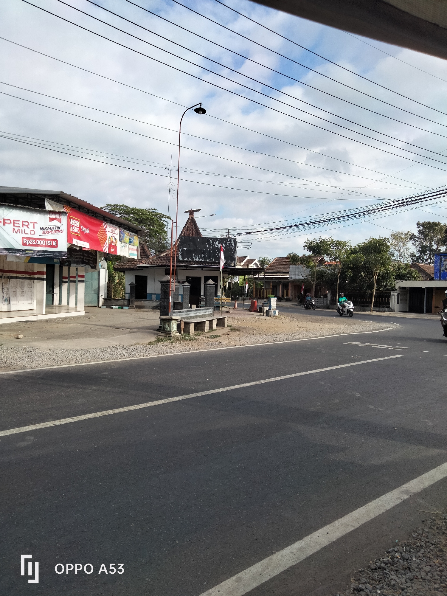 The image depicts a street scene in Wonoanti, Indonesia. A paved road with white lane markings runs diagonally across the lower portion of the frame. On the left, a building with white shutters and red and white banners is visible. One banner advertises "EXPERT MILD Rp23.000 ISI 16". Further down the street, on the right, are more buildings with tiled roofs and a flagpole. A street lamp with a dark lamp head is positioned on the left side of the road. Several utility wires crisscross the sky, which is partly cloudy. In the background, trees and residential structures can be seen. Three motorcycles are present: one is parked near the buildings on the left, another is being ridden by a person wearing a light blue shirt on the right side of the road, and a third is partially visible further down the road on the right. The overall impression is that of a typical street in a populated area with commercial establishments.