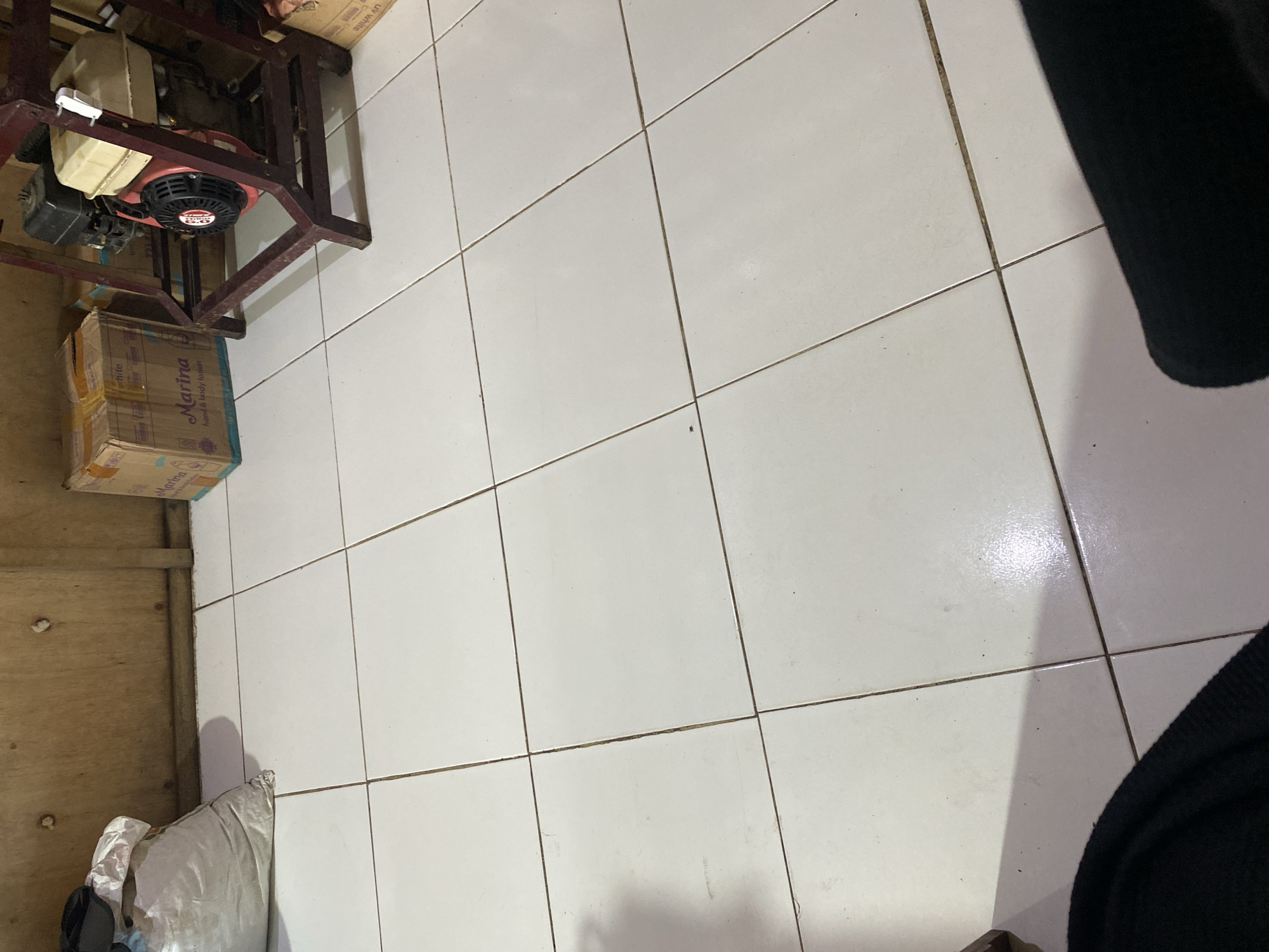 In a room with tiled flooring, a sack of unknown material rests near a wooden wall. To the right, a generator sits on a red metal frame. A cardboard box labeled "Marina hand & body lotion" is positioned to the left of the generator. The scene appears to be a storage area, possibly in Ta', Indonesia, given the context. There are no visible people in the image.