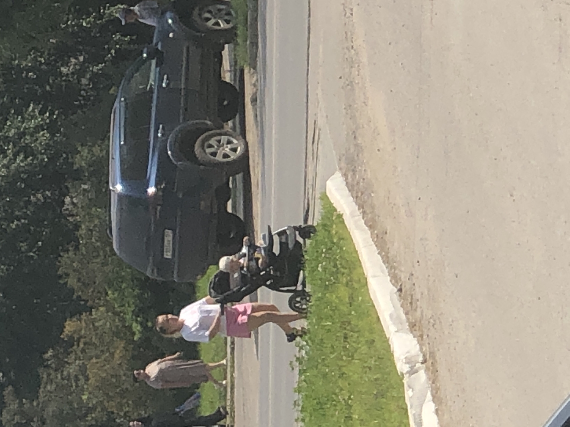 A woman wearing a white t-shirt and pink shorts pushes a stroller containing a baby. She is walking on a paved path next to a grassy area. A dark blue SUV is parked on the street behind them. In the background, there are trees and other people walking. The scene appears to be in a residential area or park. The image was taken in Solikamsk, Russia.