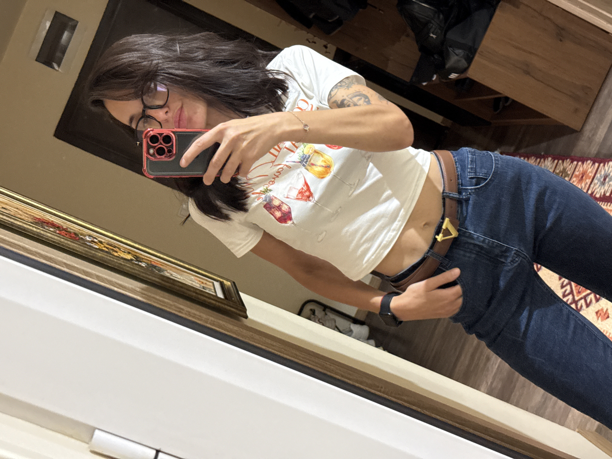 A young woman is taking a selfie in a mirror. She's wearing glasses, a white t-shirt with cocktail designs, dark blue jeans with a brown belt and a gold buckle, and a bracelet. She is holding a red-cased smartphone. The reflection shows an interior setting, including part of a wooden cabinet and a rug. The location in Pleven, Bulgaria, cannot be determined from the image.