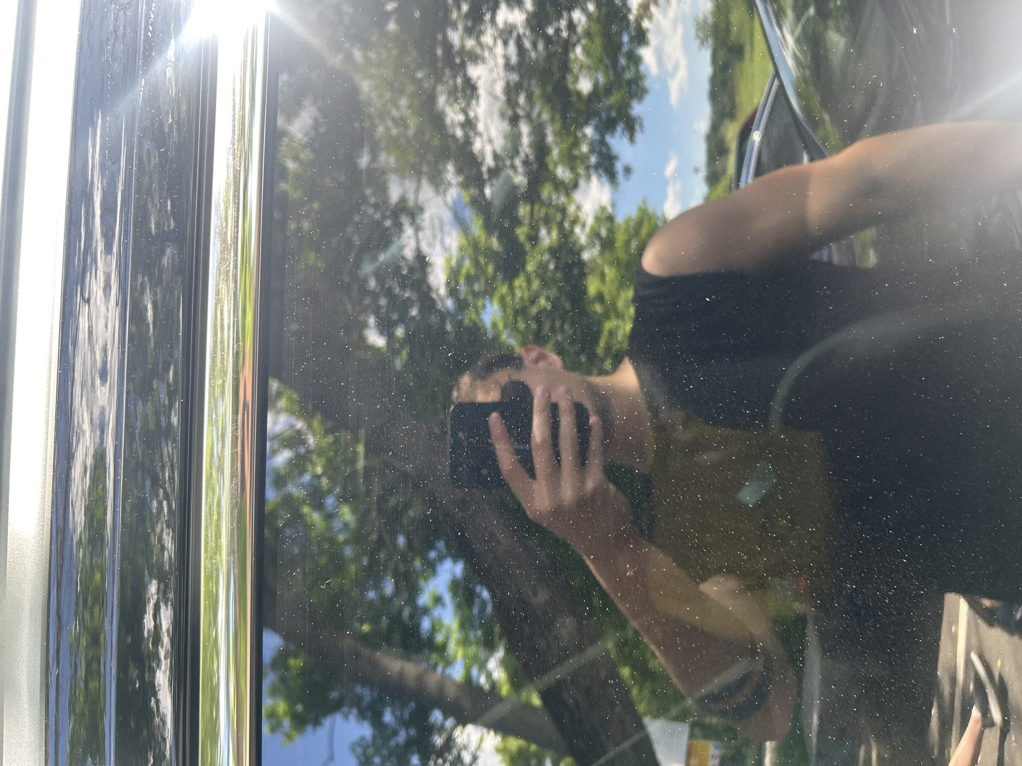 The image shows a reflection in a car door, likely outdoors. The reflection includes trees, a partly cloudy sky, and a person holding a smartphone, seemingly taking a picture of the reflection. The car's trim is visible along the left edge of the frame, featuring silver and dark-colored elements. The surface of the car door is dusty. The location in Elhovo, Bulgaria is not determinable from the visual information.