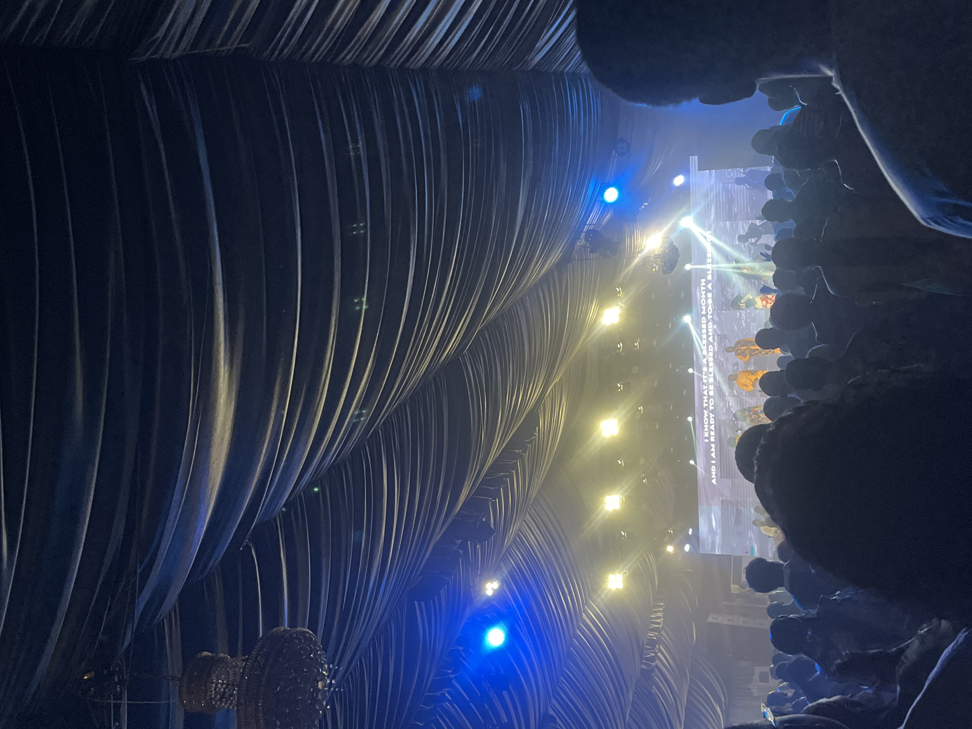 The image shows an indoor event, likely a concert or stage production, viewed from an audience perspective. The ceiling is adorned with draped, vertically-striped fabric, possibly curtains or a decorative structure. The scene is lit with a mix of yellow and blue stage lights. A large screen in the background displays an image, and text reading "I KNOW THAT IT'S A BLESSED MONTH AND I AM READY TO BE BLESSED AND-TO-BE A BLESS" is also visible. Audience members are partially visible in the foreground, mostly as blurred silhouettes and heads, due to the low-light conditions and viewing angle. The specific city of Galadimawa Village, Nigeria, is not explicitly depicted in the image.