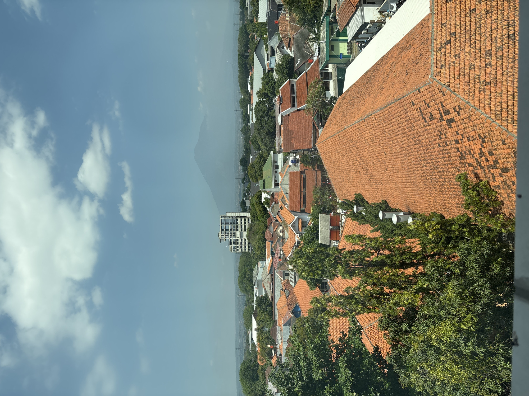 A high-angle view captures a dense urban residential area in Cirebon, Indonesia, with a prominent mountain in the distant background. The foreground is dominated by a large, reddish-orange tiled roof, its surface showing some discoloration and dark patches. To the left, lush green trees with dense foliage overhang the roof. Beyond this, a cityscape unfolds with numerous houses, most featuring similar orange tiled roofs, interspersed with patches of greenery from trees and gardens. A multi-story building with a white facade and visible windows stands out among the lower structures in the middle ground. In the distance, the hazy silhouette of a mountain rises against a pale blue sky with scattered white clouds. The overall scene suggests a warm, sunny day in a moderately populated urban environment.