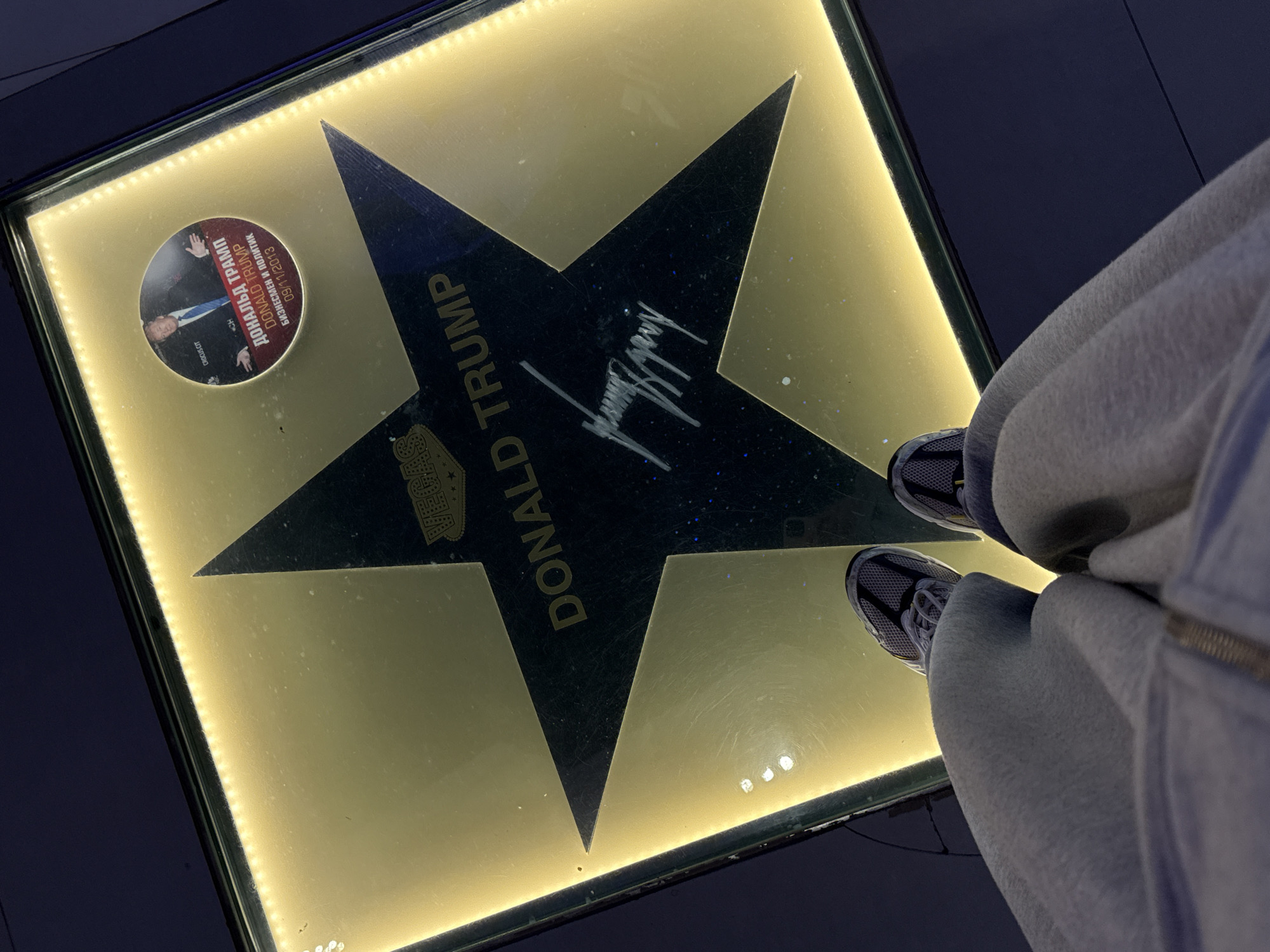 A close-up, top-down shot shows a person's feet, clad in grey sweatpants and athletic shoes, standing on a large, illuminated star on a polished floor. The star is dark with shimmering particles and has the name "DONALD TRUMP" inscribed in white letters along its left arm. A signature, also appearing to be Donald Trump's, is scrawled across the center of the star.
To the left of the star, a circular plaque is embedded in the floor. This plaque features a photograph of Donald Trump in a suit and tie, with his hands held up in a gesture. Text on the plaque, in Cyrillic and English, reads "ДОНАЛЬД ТРАМП" and "DONALD TRUMP," followed by "БИЗНЕСМЕН И ПОЛИТИК" (Businessman and Politician) and "09/11/2013." The word "HOLLYWOOD" is visible at the bottom of the plaque. The star and plaque are set within a golden, illuminated square on a dark, reflective floor. The lighting suggests an indoor environment, possibly a museum or a public walkway designed to commemorate famous individuals. The overall mood is neutral, focused on the display and the person observing it.
