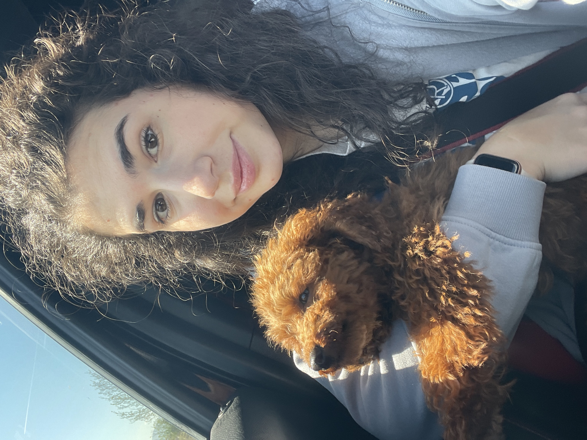 A selfie image depicts a woman with curly dark hair, light skin, and brown eyes. She is smiling slightly. She is wearing a light-colored hooded sweatshirt with a design, a dark-colored shirt underneath, and a seatbelt. The woman holds a small, reddish-brown poodle in her arms. The setting appears to be inside a car, suggested by the window and interior elements visible. The location is unknown.