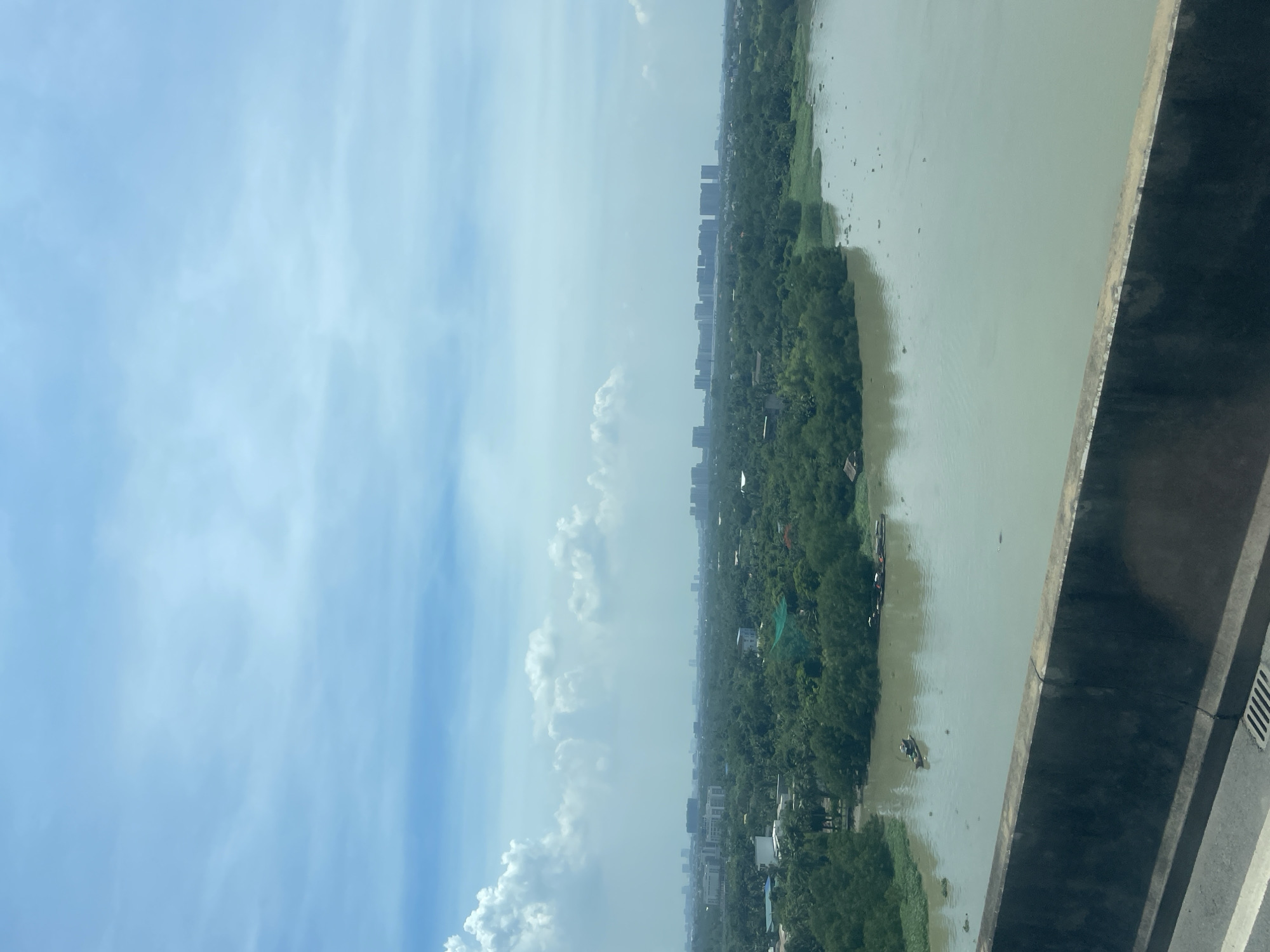 The image captures a wide, murky river flowing through a lush, green landscape in Dong Nai City, Vietnam. In the foreground, a concrete barrier marks the edge of what appears to be a bridge or elevated roadway, with a sliver of asphalt and a metal grate visible. The river itself is calm, with a few small boats, including a single, narrow vessel with a person in it, dotting its surface. The far bank is densely vegetated, with a scattering of houses and buildings interspersed among the trees. Beyond the greenery, a cityscape emerges on the horizon, characterized by a series of tall, modern buildings that contrast with the natural surroundings. The sky above is a pale blue, with scattered white clouds, suggesting a bright but possibly hazy day. The overall impression is one of a developing urban area coexisting with its natural environment.